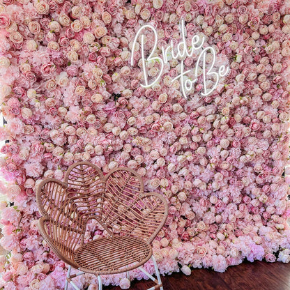 A stunning flower wall filled with an abundance of pink roses. In the center, neon lights spell out &quot;Bride To Be&quot;. A wicker chair is placed in front, creating a romantic bridal setting.