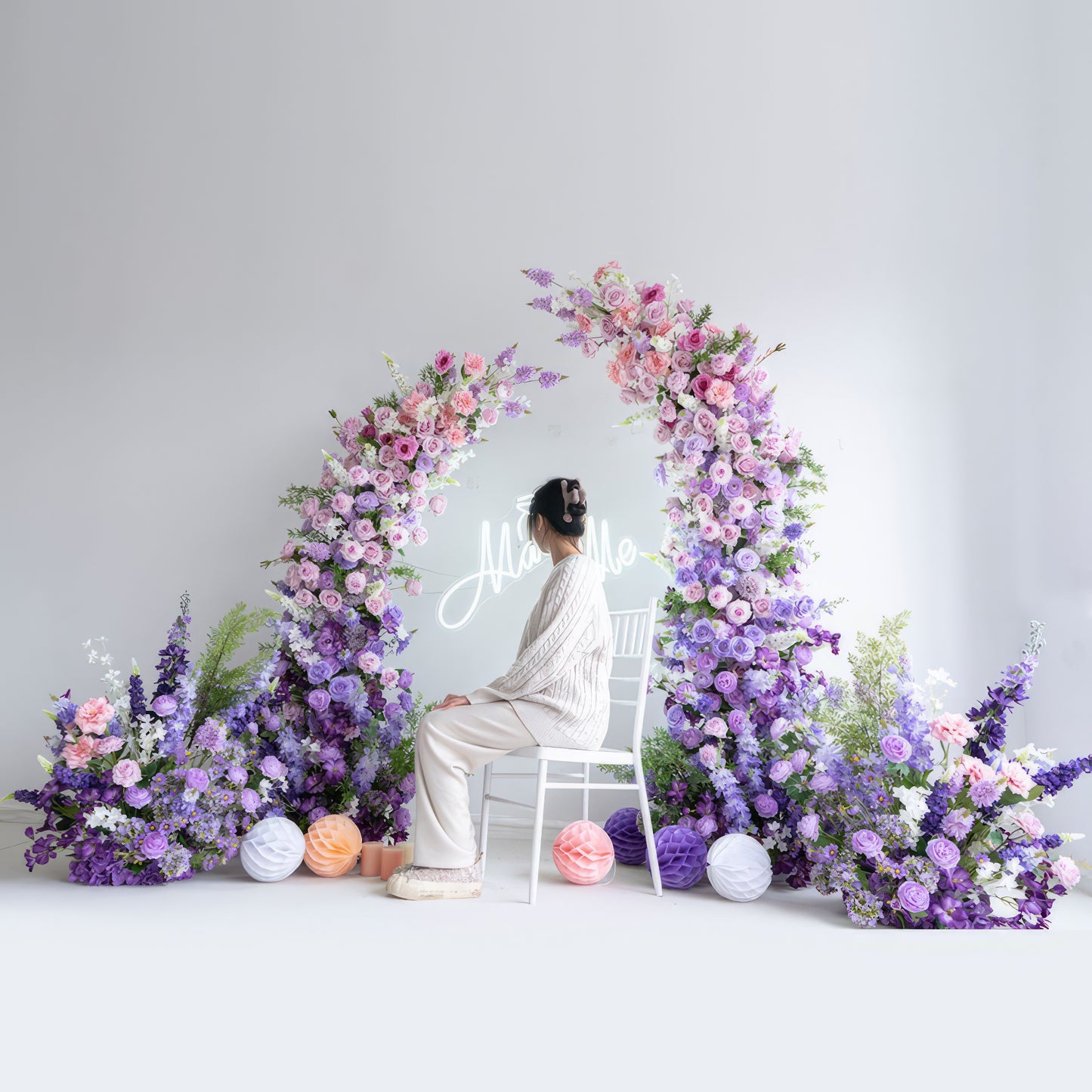 This romantic floral arch from Valar Flowers features a soft pastel palette of blush pink, lavender, and white blooms—including roses, hydrangeas, and delphiniums—arranged in two cascading columns. A glowing "Marry Me" neon sign hangs at its center, with a woman seated on a white chair in front, creating a dreamy, intimate atmosphere ideal for a heartfelt proposal or engagement photoshoot.
