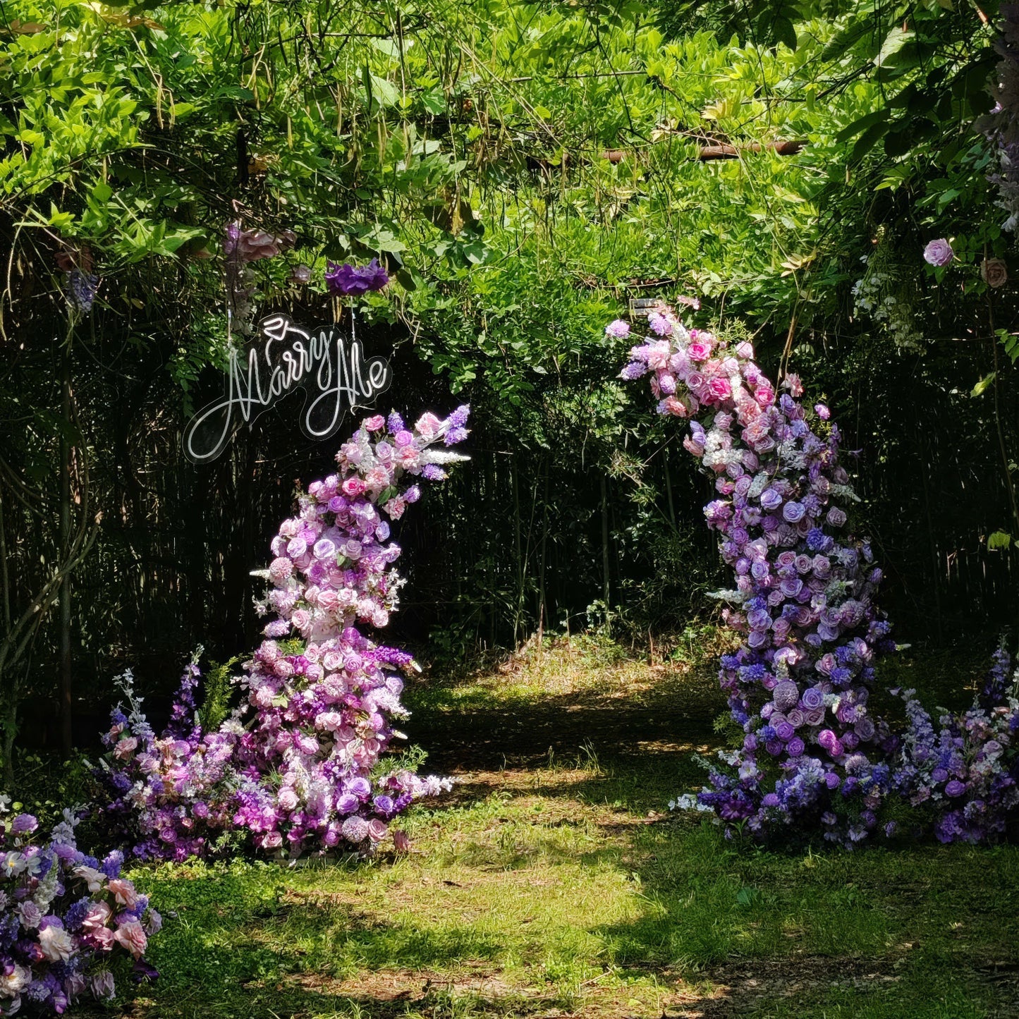 This romantic floral arch from **Valar Flowers** is a lush tapestry of blush pink, lavender, and white blooms—including roses, peonies, and hydrangeas. Suspended at its center is a glowing "Marry Me" neon sign. Set in a sun-dappled, leafy garden, the design evokes a dreamy, intimate atmosphere ideal for a heartfelt proposal or engagement photoshoot.