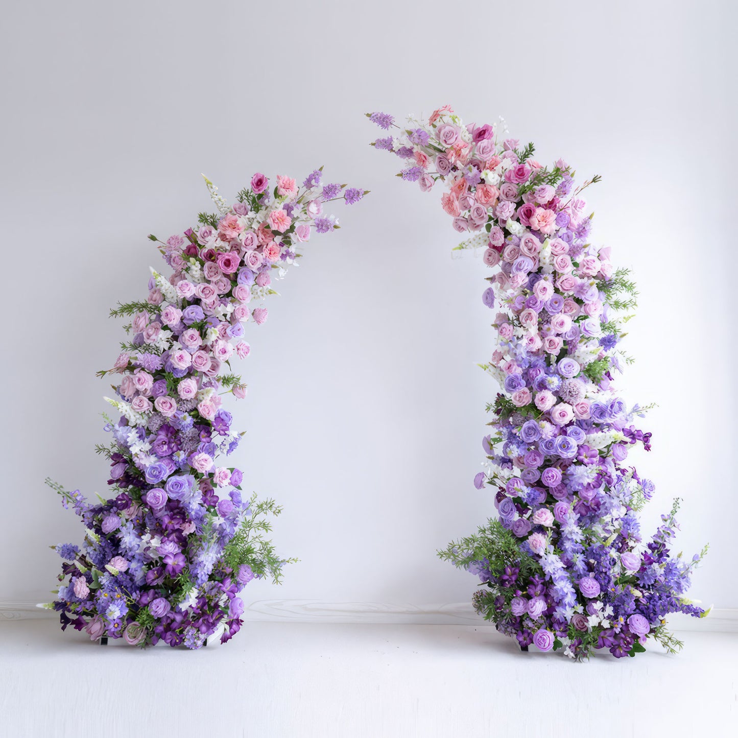 This elegant floral arch from Valar Flowers features a soft gradient of blush pink, lavender, and deep purple blooms—including roses, ranunculus, and hydrangeas—arranged in two cascading columns. Set against a minimalist white backdrop, the design evokes a romantic, sophisticated atmosphere ideal for weddings, engagement photoshoots, or elegant celebrations.
