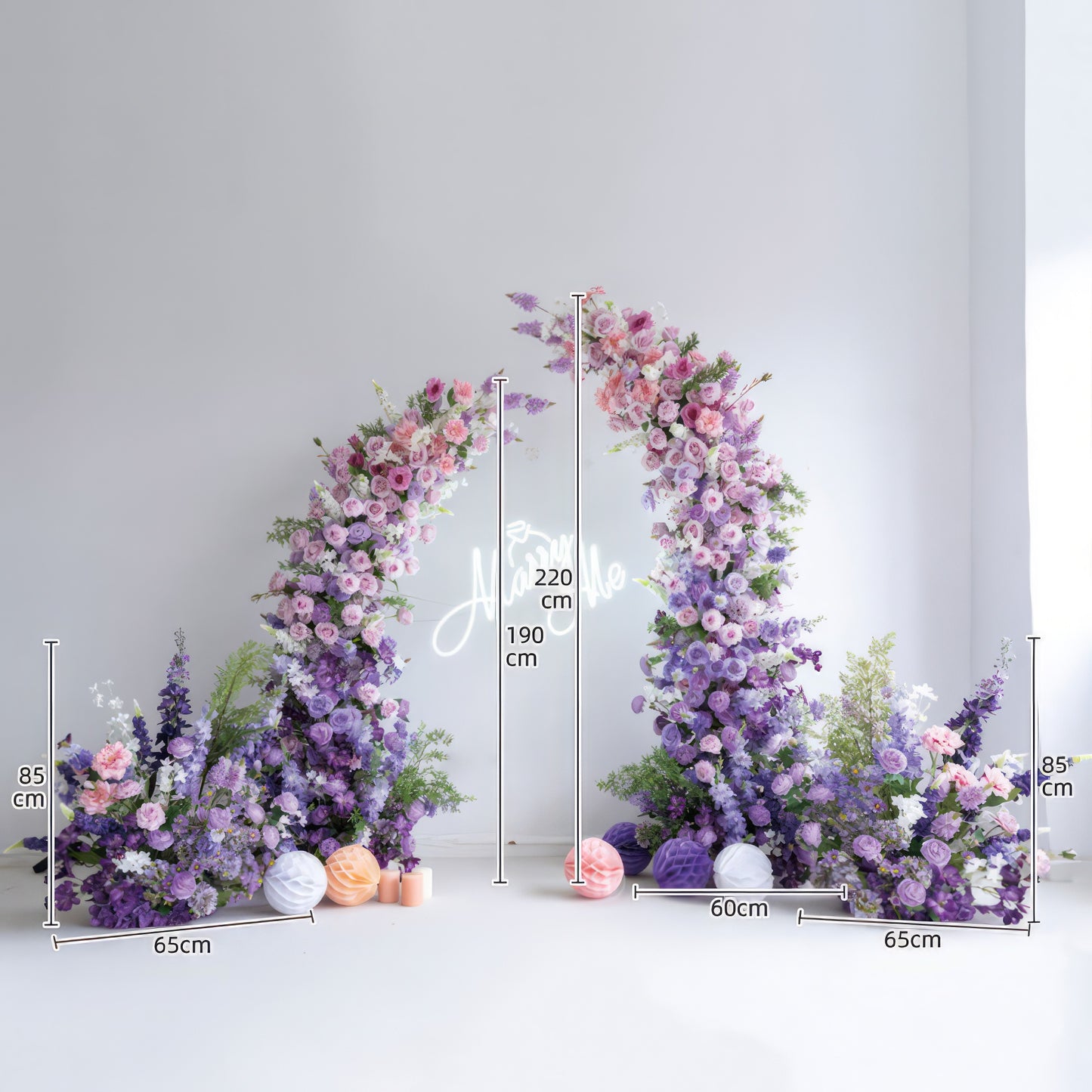 This elegant floral arch from Valar Flowers features a soft gradient of blush pink, lavender, and deep purple blooms—including roses, ranunculus, and hydrangeas—arranged in two cascading columns. A glowing "Marry Me" neon sign hangs at its center, with decorative paper lanterns and candles at the base. Set against a minimalist white backdrop, the design evokes a romantic, sophisticated atmosphere ideal for weddings, engagement photoshoots, or elegant celebrations.