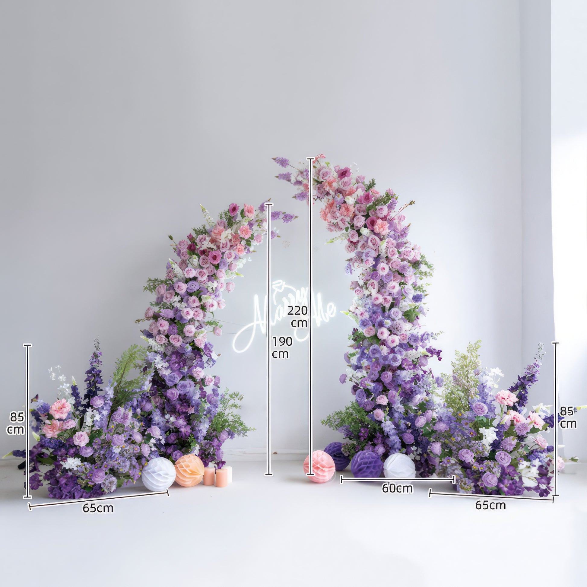 This elegant floral arch from Valar Flowers features a soft gradient of blush pink, lavender, and deep purple blooms—including roses, ranunculus, and hydrangeas—arranged in two cascading columns. A glowing "Marry Me" neon sign hangs at its center, with decorative paper lanterns and candles at the base. Set against a minimalist white backdrop, the design evokes a romantic, sophisticated atmosphere ideal for weddings, engagement photoshoots, or elegant celebrations.