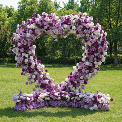 This romantic heart-shaped floral arch from **Valar Flowers** is a lush tapestry of deep purple, soft lavender, and crisp white blooms—including roses, peonies, and delicate filler flowers. Set on a sunlit green lawn framed by leafy trees, the dense, layered design creates a rich, three-dimensional texture, evoking a dreamy, romantic atmosphere ideal for a heartfelt proposal, engagement photoshoot, or elegant outdoor celebration.