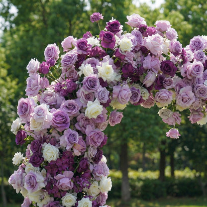 This close-up of a romantic floral arch from **Valar Flowers** showcases a lush tapestry of deep purple, soft lavender, and crisp white blooms—including roses, peonies, and delicate filler flowers. The dense, layered design creates a rich, three-dimensional texture, evoking a dreamy, romantic atmosphere ideal for a heartfelt proposal, engagement photoshoot, or elegant outdoor celebration. Set against a sunlit green lawn and leafy trees, it exudes a soft, joyful ambiance.