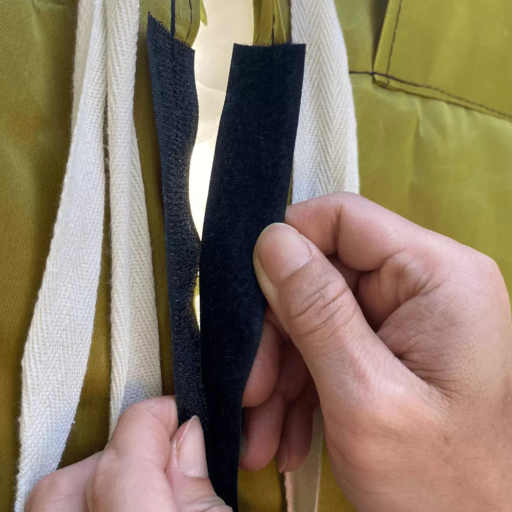 This close - up photo shows two hands separating a piece of black hook - and - loop fastener (Velcro). Surrounding it are some white fabric straps against a mustard - colored fabric background. The image focuses on the action of opening the fastener. 