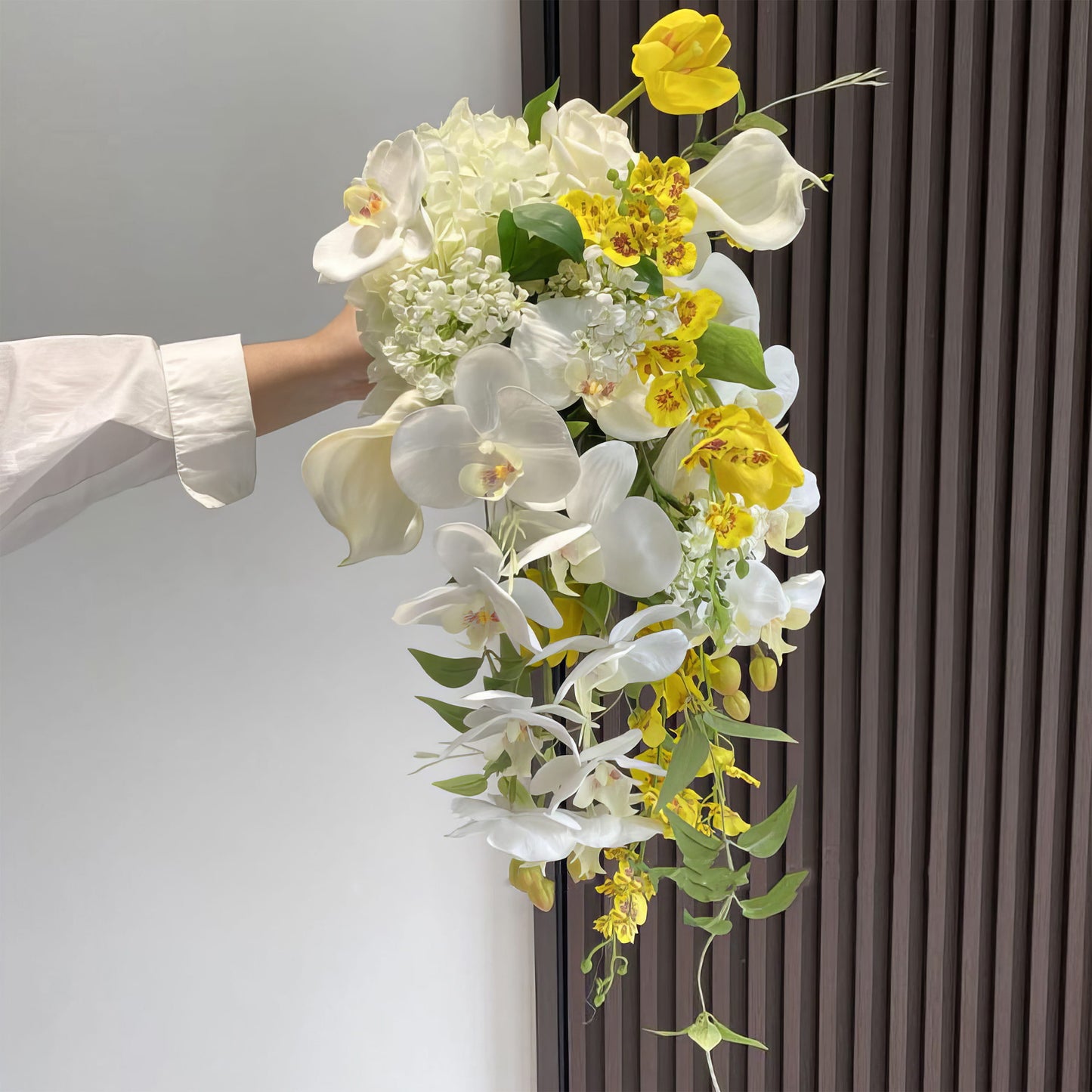 This cascading bridal bouquet blends white orchids, calla lilies, hydrangeas, and delicate white blooms with vibrant yellow flowers and green leaves. The flowing design creates a romantic, elegant look, perfect for weddings, showcasing a harmonious mix of colors and textures. 