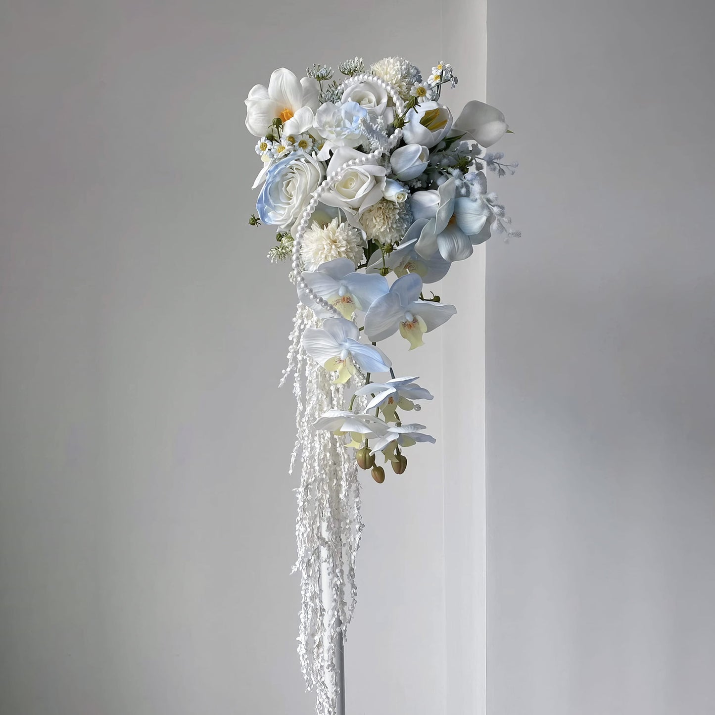This cascading bridal bouquet features a dreamy mix of white and light blue blooms—roses, orchids, calla lilies, and tiny daisies. Adorned with pearl strands and trailing white vines, its flowing design exudes elegance, perfect for weddings, symbolizing romance and new beginnings. 