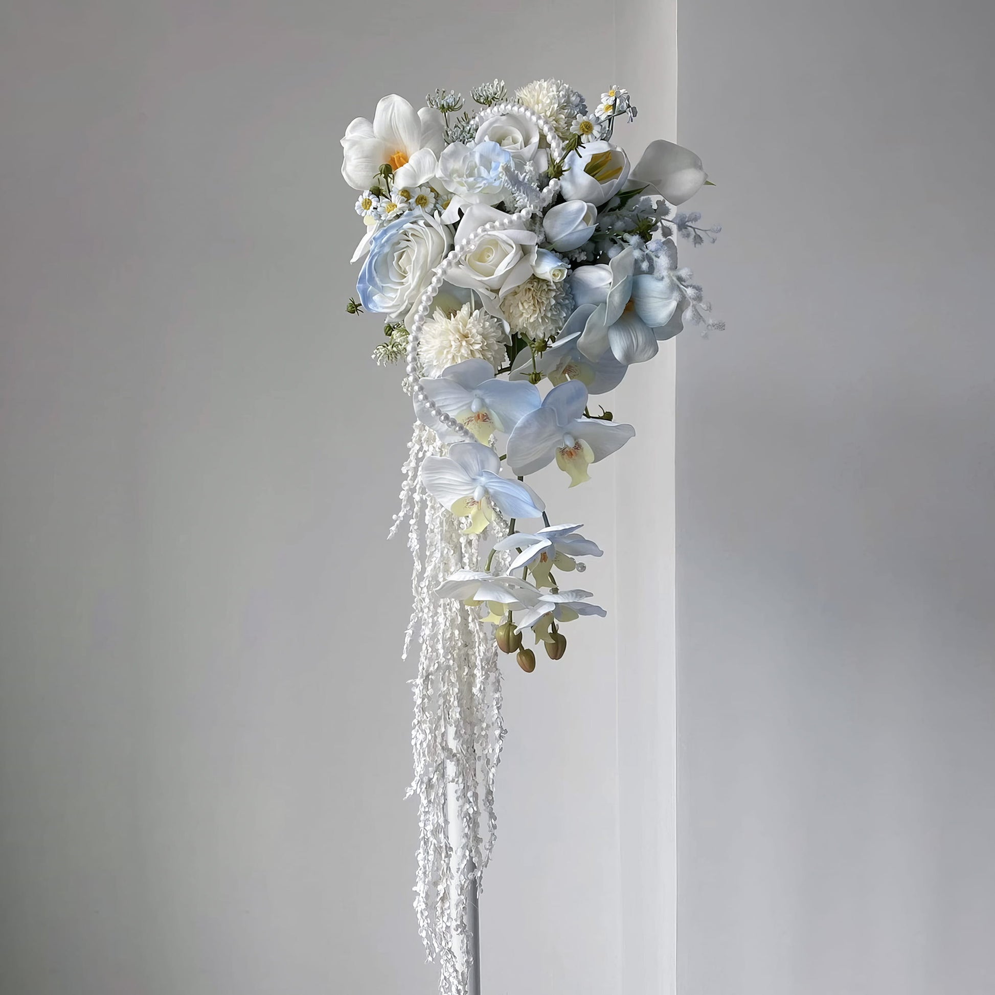 This cascading bridal bouquet features a dreamy mix of white and light blue blooms—roses, orchids, calla lilies, and tiny daisies. Adorned with pearl strands and trailing white vines, its flowing design exudes elegance, perfect for weddings, symbolizing romance and new beginnings. 