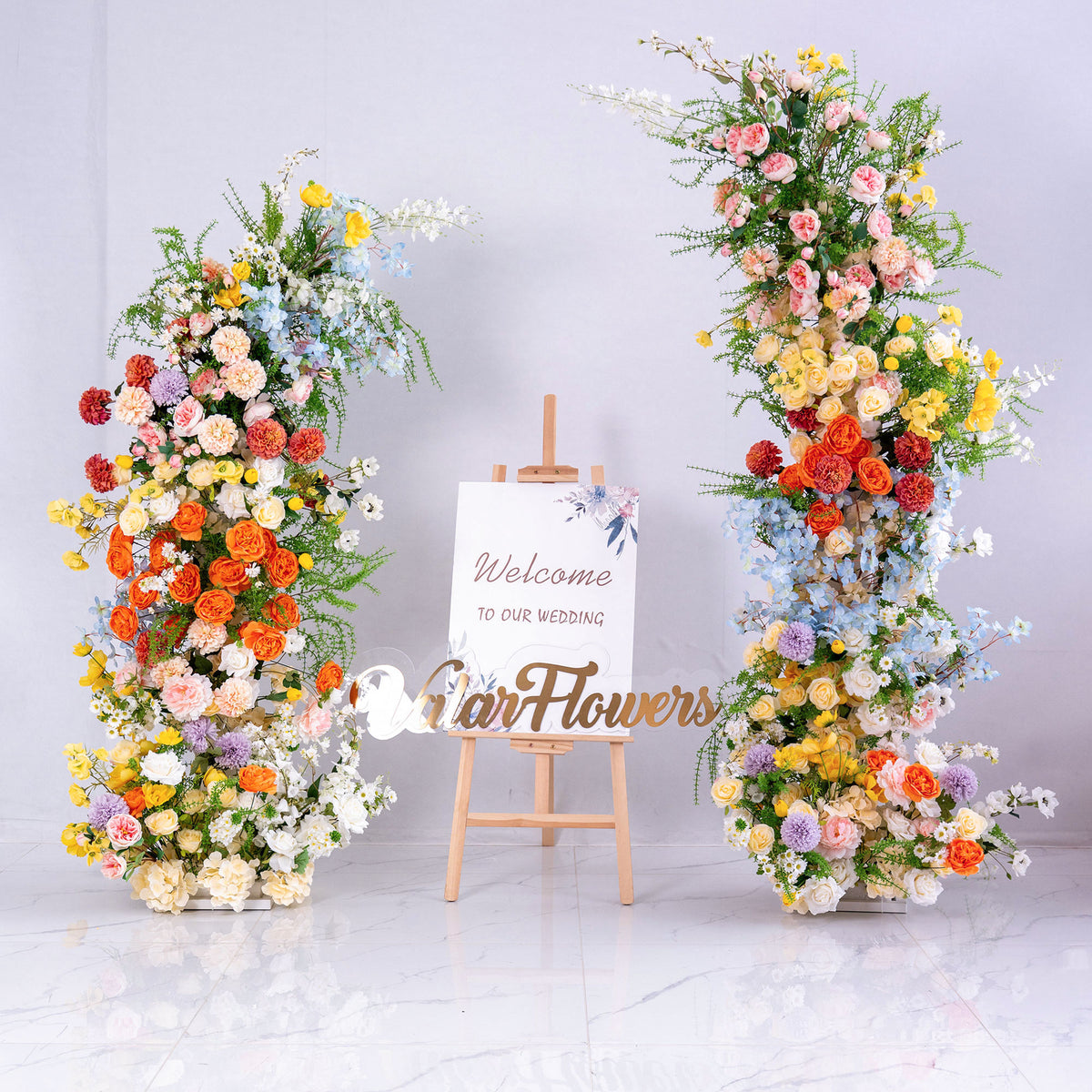 This photo shows a wedding - themed setup. Two large floral installations flank a sign saying “Welcome to Our Wedding.” Flowers include roses, peonies, chrysanthemums, and delphinium, in vibrant colors like orange, yellow, pink, and blue. The lush, colorful blooms create a cheerful, romantic vibe, perfect for greeting wedding guests and setting an elegant, festive tone for the celebration. 