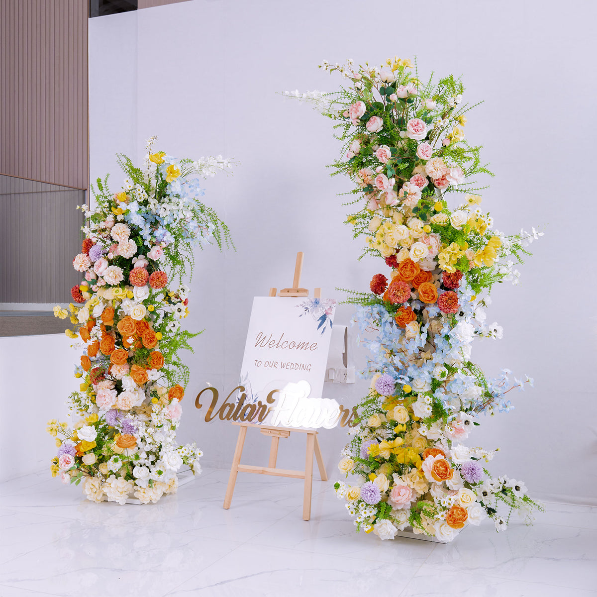 This wedding - themed setup has two tall floral installations flanking a “Welcome to Our Wedding” sign. Flowers like roses, peonies, chrysanthemums, and delphinium come in vibrant colors—orange, yellow, pink, blue. The lush, colorful blooms create a cheerful, romantic vibe, perfect for greeting guests and setting an elegant, festive tone for the celebration. 