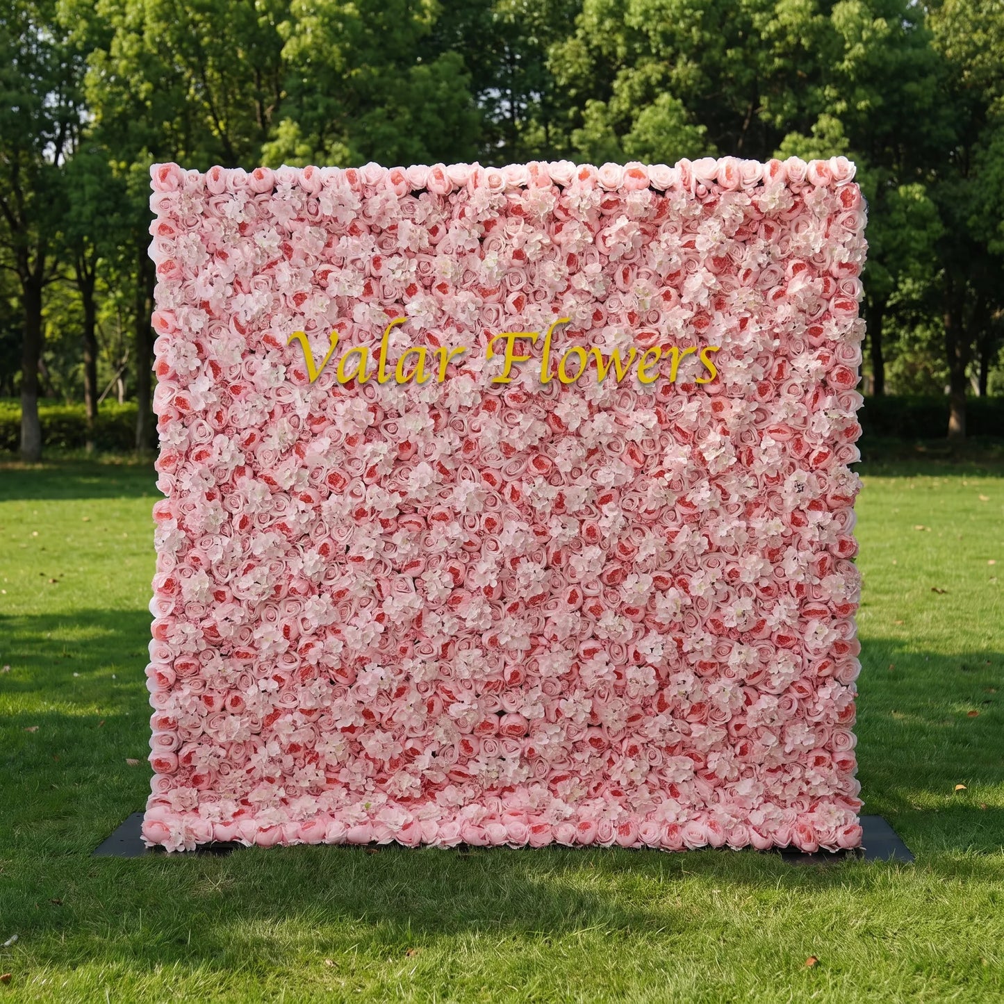 Dense pastel pink artificial flower wall filled with soft blush roses and fluffy white hydrangeas, set on a sunlit green lawn with leafy trees behind. Centered glossy golden "Valar Flowers" signage adds custom branding appeal. This reusable zero-maintenance floral backdrop suits wedding photo booths, bridal parties, boutique shop displays and pink-themed social media shoots, offering full lush coverage for romantic, eye-catching event decor.