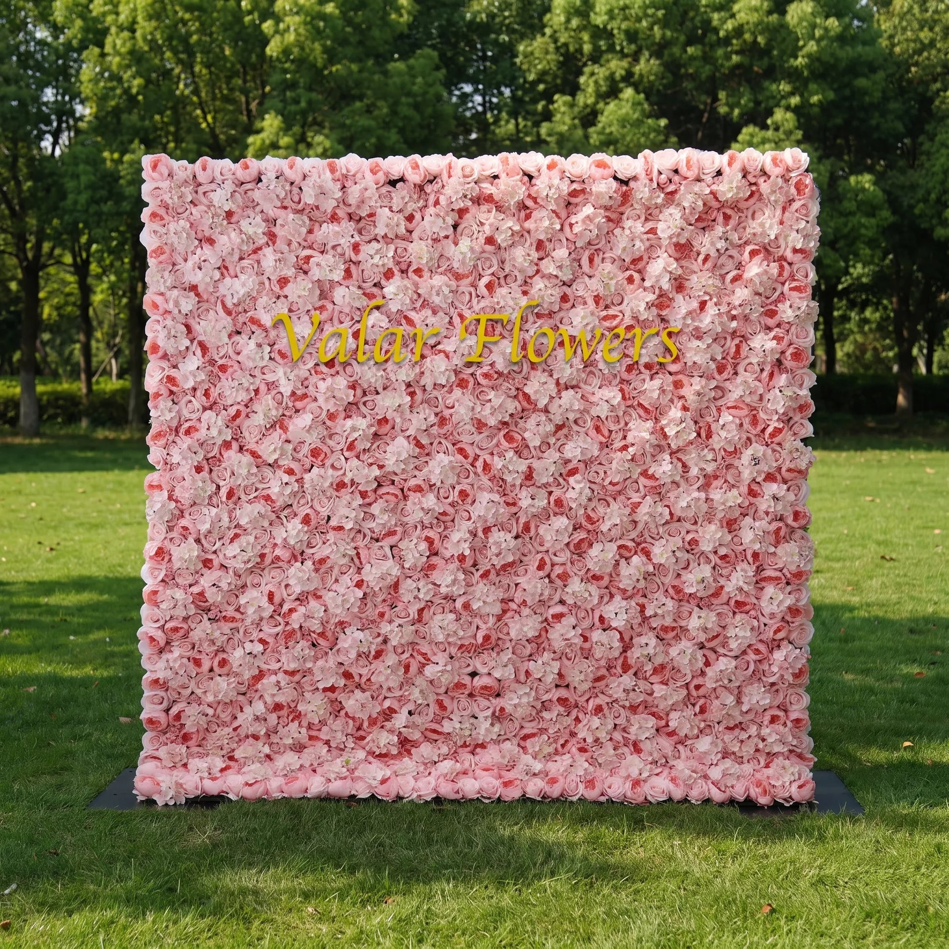 Dense pastel pink artificial flower wall filled with soft blush roses and fluffy white hydrangeas, set on a sunlit green lawn with leafy trees behind. Centered glossy golden "Valar Flowers" signage adds custom branding appeal. This reusable zero-maintenance floral backdrop suits wedding photo booths, bridal parties, boutique shop displays and pink-themed social media shoots, offering full lush coverage for romantic, eye-catching event decor.