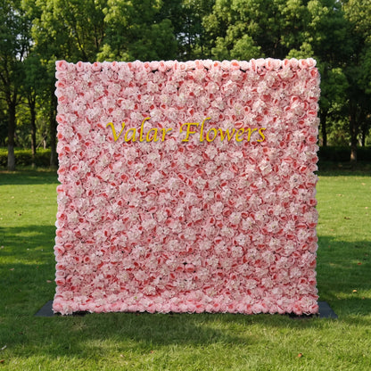 Dense pastel pink artificial flower wall filled with soft blush roses and fluffy white hydrangeas, set on a sunlit green lawn with leafy trees behind. Centered glossy golden "Valar Flowers" signage adds custom branding appeal. This reusable zero-maintenance floral backdrop suits wedding photo booths, bridal parties, boutique shop displays and pink-themed social media shoots, offering full lush coverage for romantic, eye-catching event decor.
