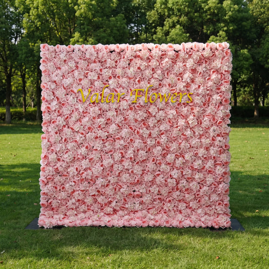Dense pastel pink artificial flower wall filled with soft blush roses and fluffy white hydrangeas, set on a sunlit green lawn with leafy trees behind. Centered glossy golden "Valar Flowers" signage adds custom branding appeal. This reusable zero-maintenance floral backdrop suits wedding photo booths, bridal parties, boutique shop displays and pink-themed social media shoots, offering full lush coverage for romantic, eye-catching event decor.