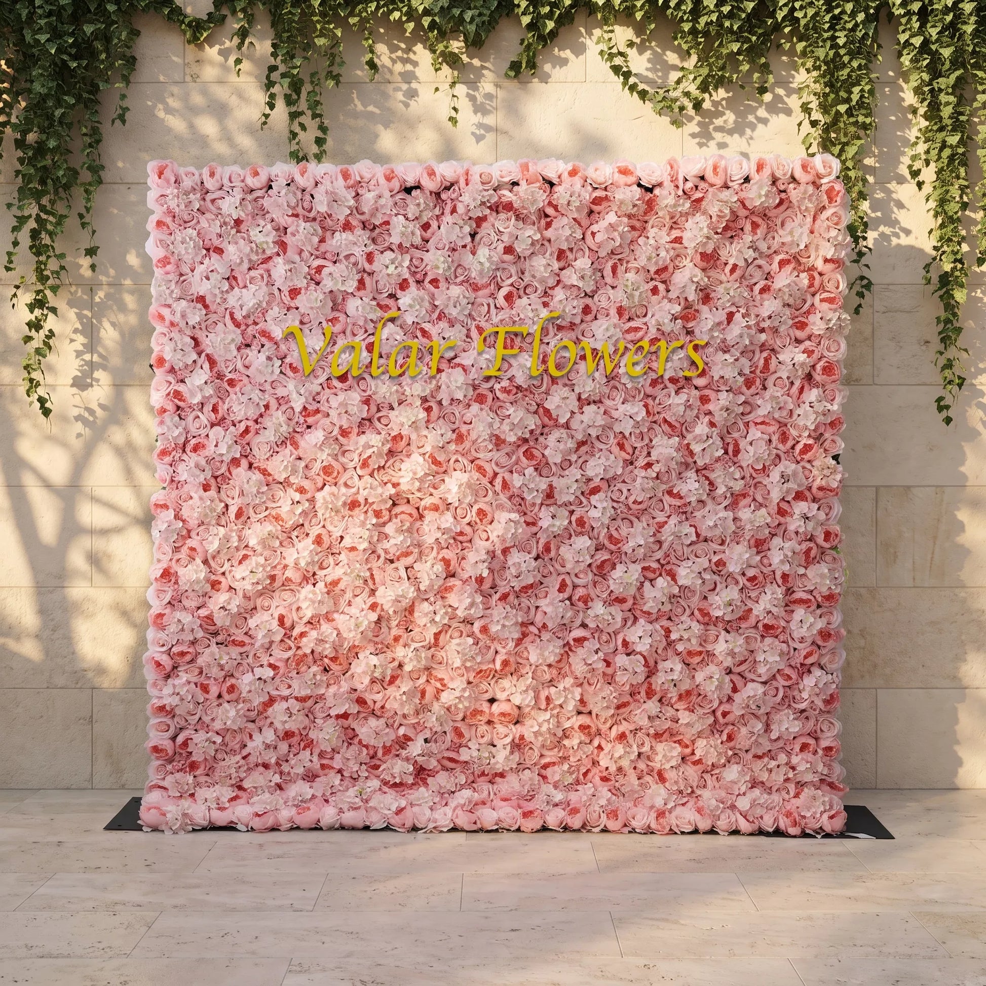 Dense blush pink artificial flower wall layered with soft roses and white hydrangeas, mounted against a stone wall draped with trailing ivy. Bright golden "Valar Flowers" custom branding sits front and center. This reusable, low-maintenance floral backdrop delivers romantic pastel texture, perfect for wedding photo booths, bridal events, boutique store displays and elegant social media content shoots for soft pink-themed celebrations.