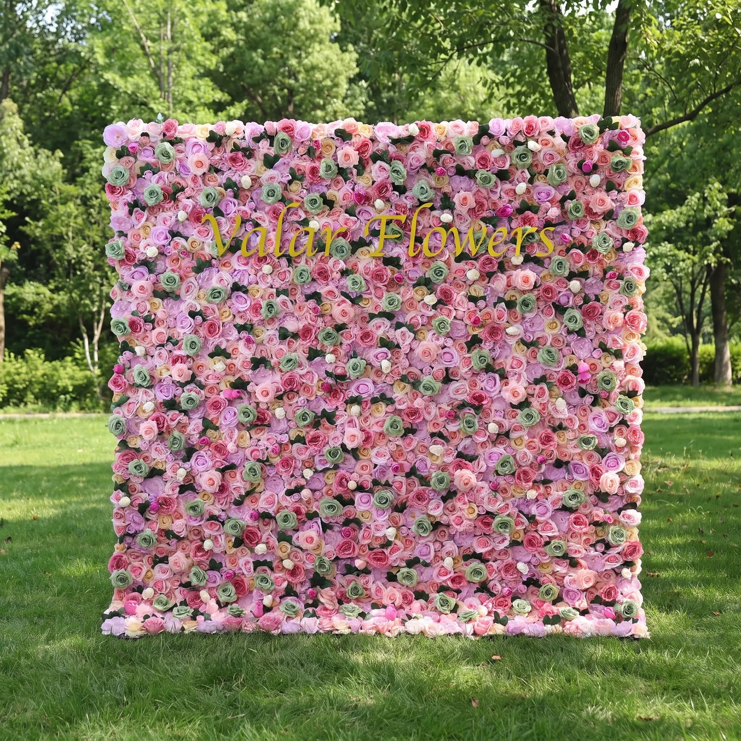 Vibrant mixed pastel artificial flower wall filled with blush, lavender, magenta roses and mint green floral buds, set on a sun-dappled green lawn. Centered glossy golden "Valar Flowers" custom branding highlights the panel. This reusable, zero-maintenance floral backdrop features full dense blooms, ideal for wedding photo booths, bridal showers, boutique decor and colorful pastel social media content shoots for whimsical event styling.