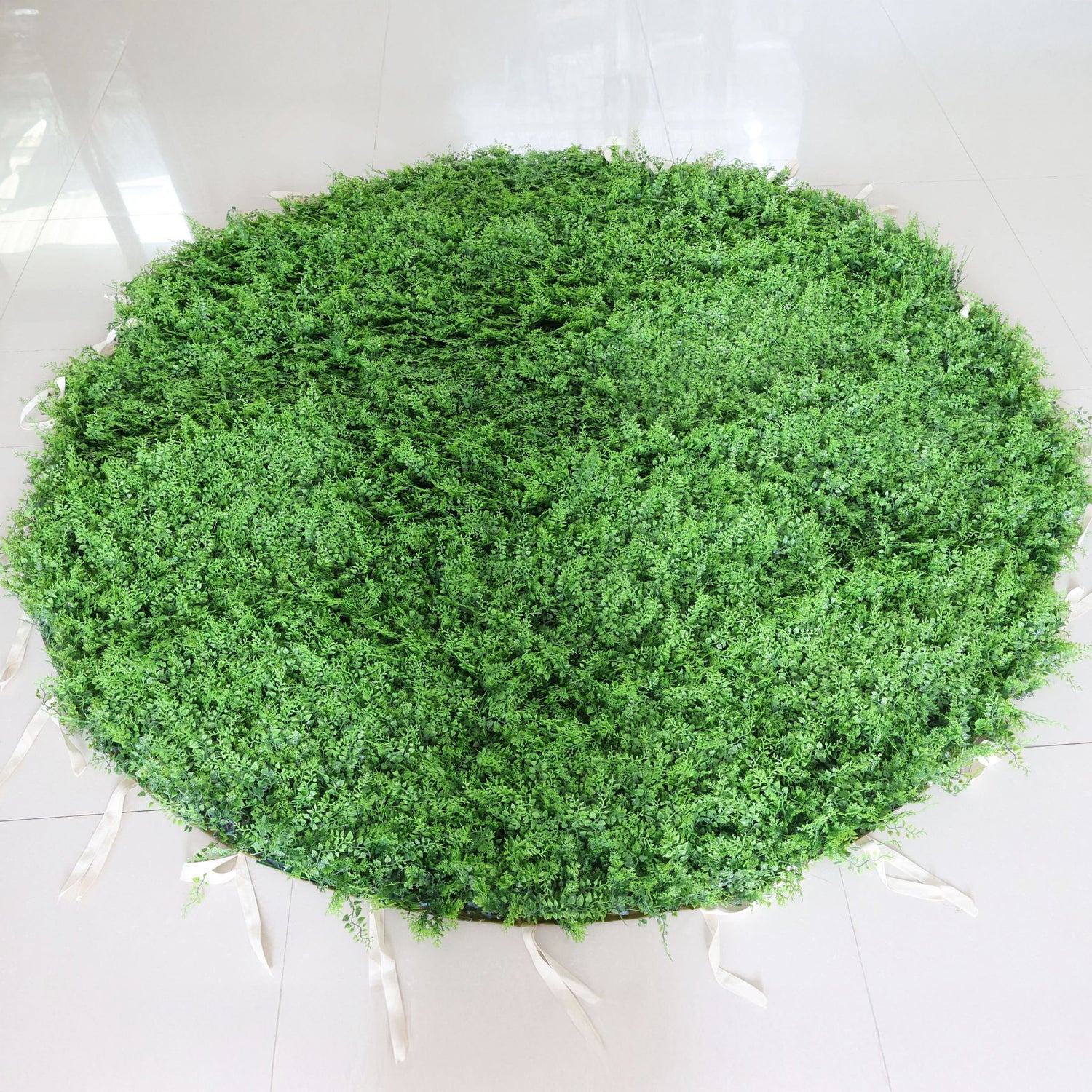 This image showcases a round artificial greenery mat by Valar Flowers, placed on a glossy tiled floor. It features dense, lush artificial foliage in a rich green hue, creating a natural and vibrant display. With attached white straps for easy handling, this piece is perfect for adding a touch of organic charm to events or spaces, exuding a fresh and sophisticated atmosphere.