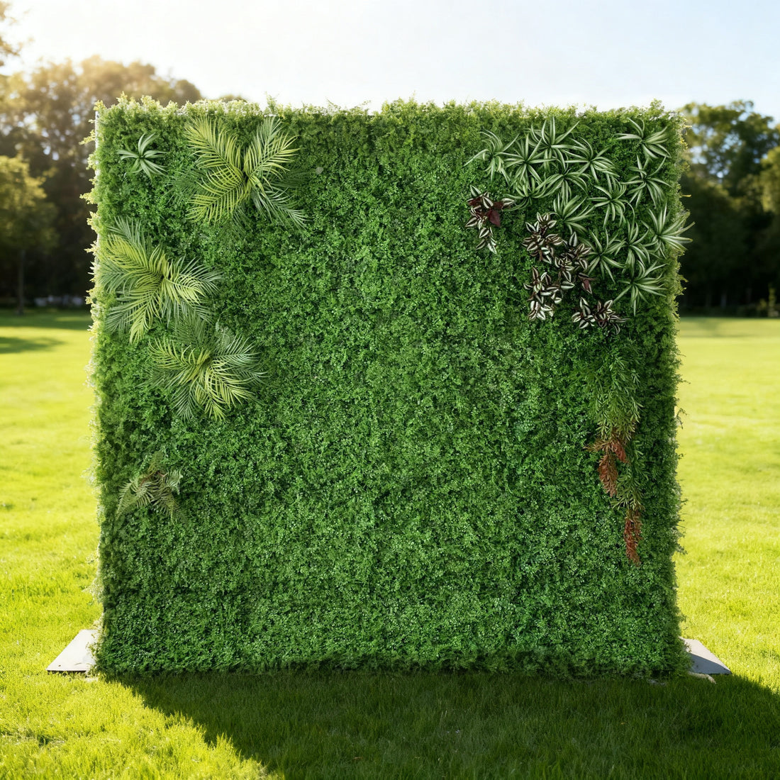 This image showcases a lush artificial greenery wall by ValarFlowers, set outdoors on a vibrant lawn. It features a dense base of green foliage, artfully accented with varied plants like palm fronds, variegated leaves, and textured greens. Bathed in sunlight, this piece blends natural diversity and realistic textures, perfect for adding organic elegance to events, exuding a serene and sophisticated atmosphere.
