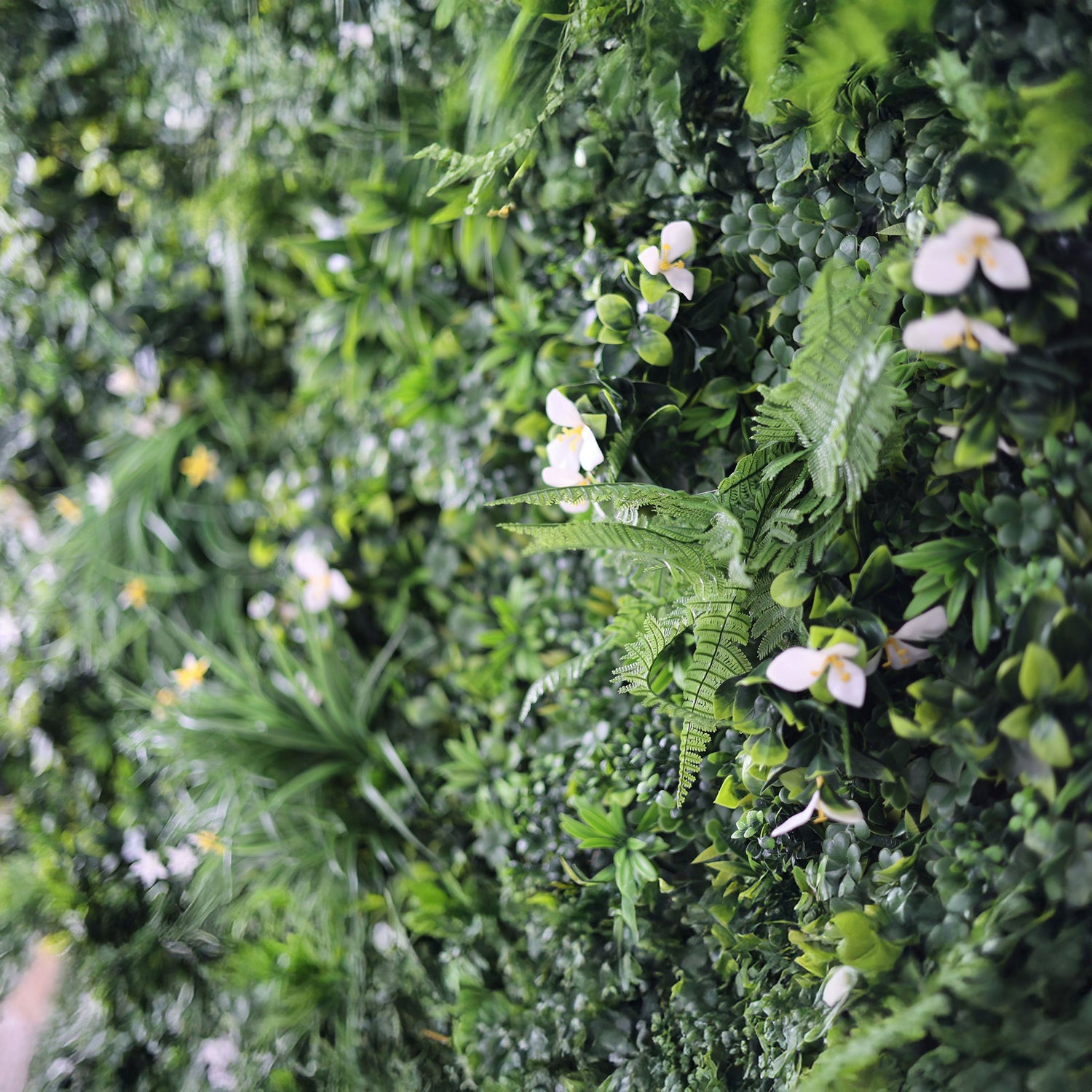 This image showcases a lush artificial greenery wall by ValarFlowers. It features a dense arrangement of varied green foliage, including ferns and textured leaves, interspersed with delicate white blooms and subtle yellow accents. The realistic textures and diverse plants create a rich, natural, and visually captivating display, perfect for adding organic elegance to events or spaces, exuding a serene and sophisticated atmosphere.