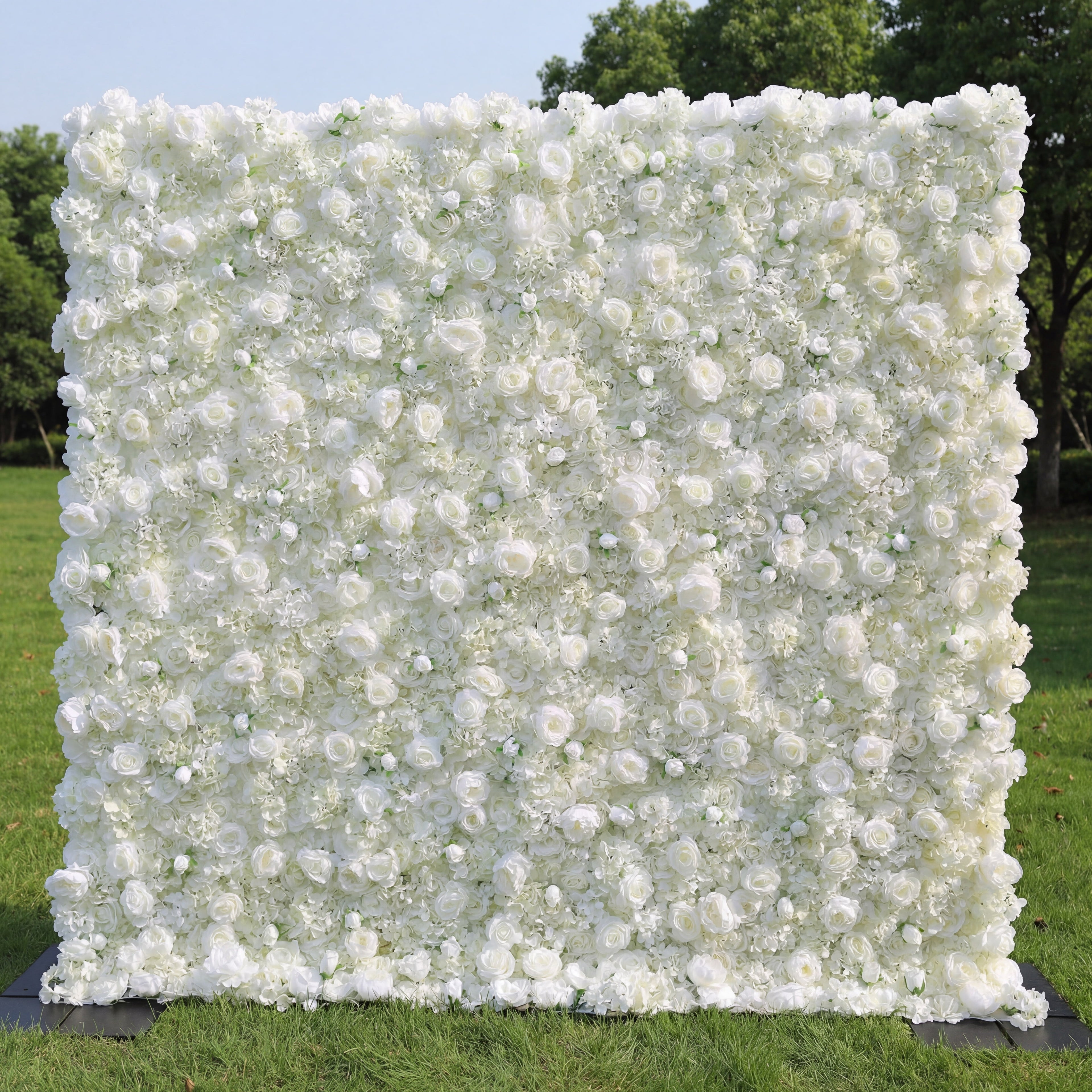 This elegant outdoor backdrop features a dense, wall-to-wall arrangement of artificial white roses and delicate filler blooms, creating a lush, velvety texture. Standing on a sunlit green lawn framed by trees, its pure, monochromatic design exudes timeless sophistication and romance, making it a stunning focal point for weddings, engagement photos, or upscale events.