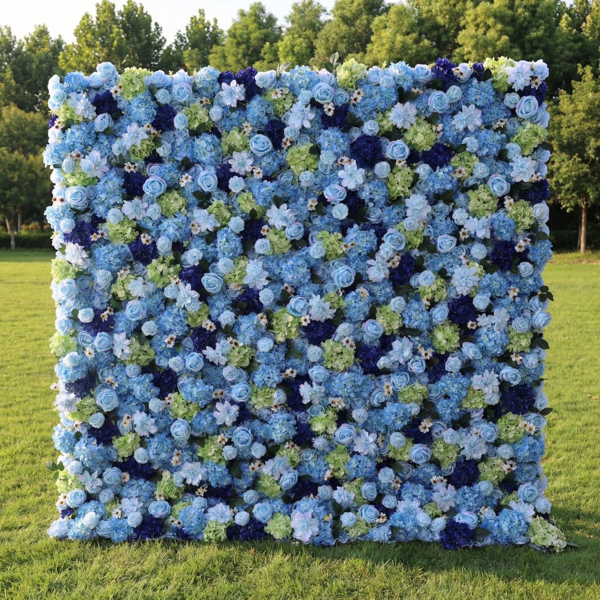 This outdoor floral backdrop stands on a lush green lawn, framed by leafy trees. It is a dense, textured wall of blooms in varying shades of blue—from soft sky-blue roses and hydrangeas to deep navy accents—interspersed with fresh green foliage. The multi-tonal, ocean-inspired palette creates a serene, dreamy atmosphere, making it ideal for garden weddings, coastal-themed events, or photo booths seeking a cool, elegant focal point.