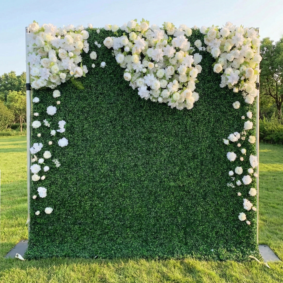 This outdoor floral backdrop from **Valar Flowers** features a lush, deep green foliage base, adorned with a cascading arrangement of white and cream blooms—including roses, hydrangeas, and delicate filler flowers. The blooms form a soft, heart-shaped design at the top, with individual flowers trailing down the sides. Set on a sunlit green lawn framed by leafy trees, the design evokes a fresh, romantic, and naturally sophisticated atmosphere ideal for weddings, photo backdrops, or elegant outdoor celebratio