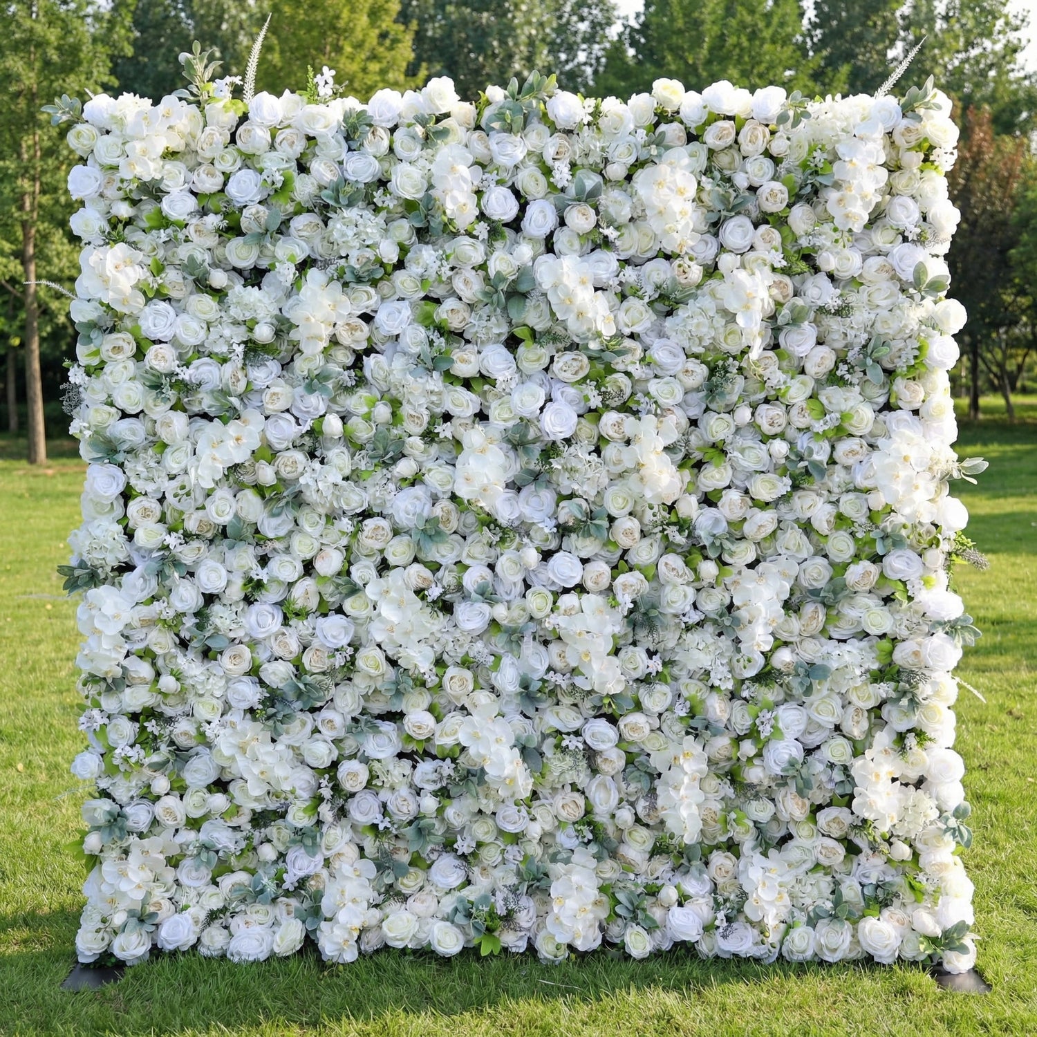 This stunning outdoor floral wall from **Valar Flowers** is a lush, all-white tapestry of blooms—including roses, hydrangeas, and delicate filler flowers—interwoven with vibrant green eucalyptus leaves. Set on a sunlit green lawn framed by leafy trees, the dense, layered design creates a rich, three-dimensional texture, evoking a fresh, romantic, and naturally elegant atmosphere ideal for weddings, photo backdrops, or elegant outdoor celebrations.