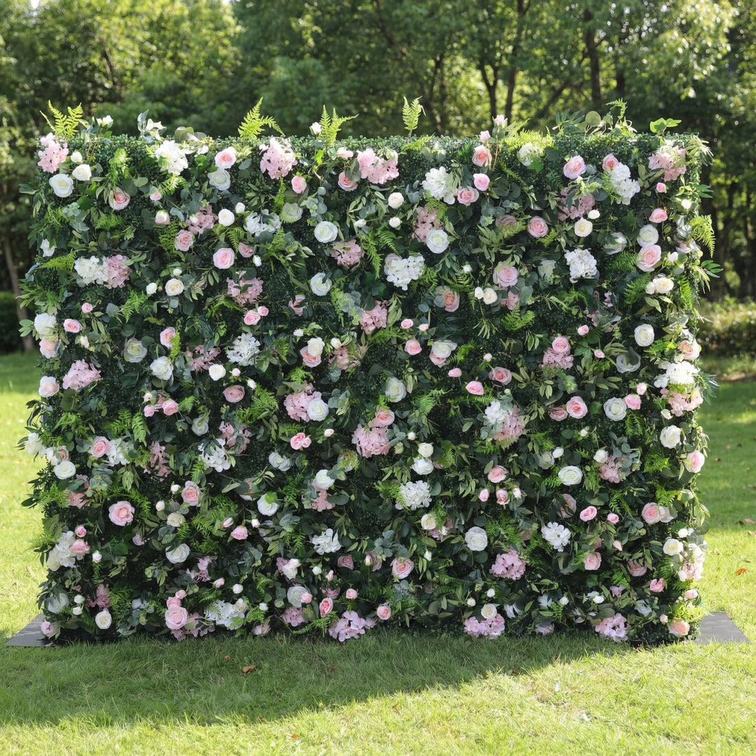 This lush outdoor floral wall is a dense, textured backdrop of deep green foliage, interspersed with a romantic mix of blush pink, soft white, and pale pink blooms—including roses, hydrangeas, and delicate filler flowers. Set on a sunlit green lawn framed by leafy trees, the layered design creates a rich, three-dimensional texture, evoking a fresh, natural, and romantic atmosphere ideal for weddings, photo backdrops, or elegant celebrations.