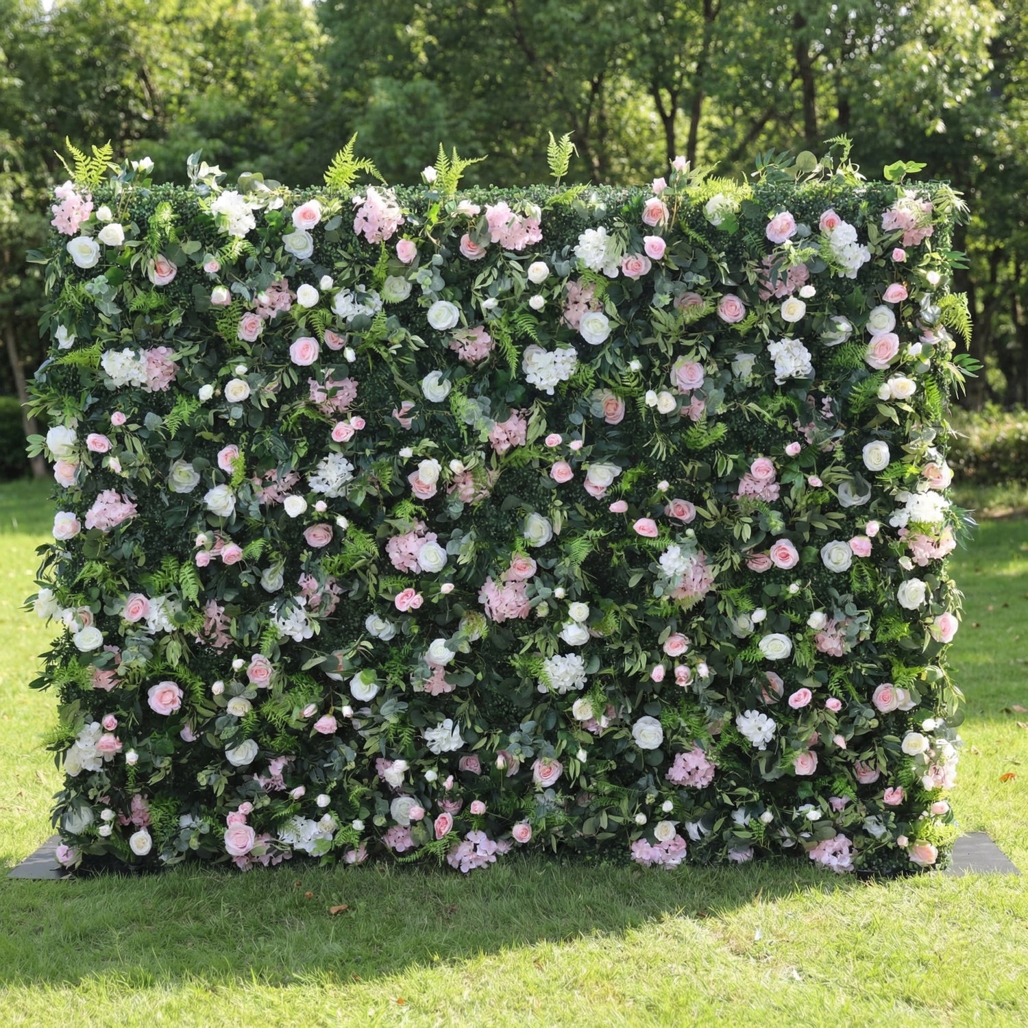 This lush outdoor floral wall is a dense, textured backdrop of deep green foliage, interspersed with a romantic mix of blush pink, soft white, and pale pink blooms—including roses, hydrangeas, and delicate filler flowers. Set on a sunlit green lawn framed by leafy trees, the layered design creates a rich, three-dimensional texture, evoking a fresh, natural, and romantic atmosphere ideal for weddings, photo backdrops, or elegant celebrations.