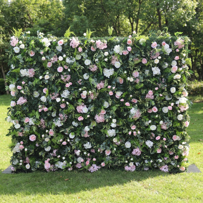 This lush outdoor floral wall is a dense, textured backdrop of deep green foliage, interspersed with a romantic mix of blush pink, soft white, and pale pink blooms—including roses, hydrangeas, and delicate filler flowers. Set on a sunlit green lawn framed by leafy trees, the layered design creates a rich, three-dimensional texture, evoking a fresh, natural, and romantic atmosphere ideal for weddings, photo backdrops, or elegant celebrations.