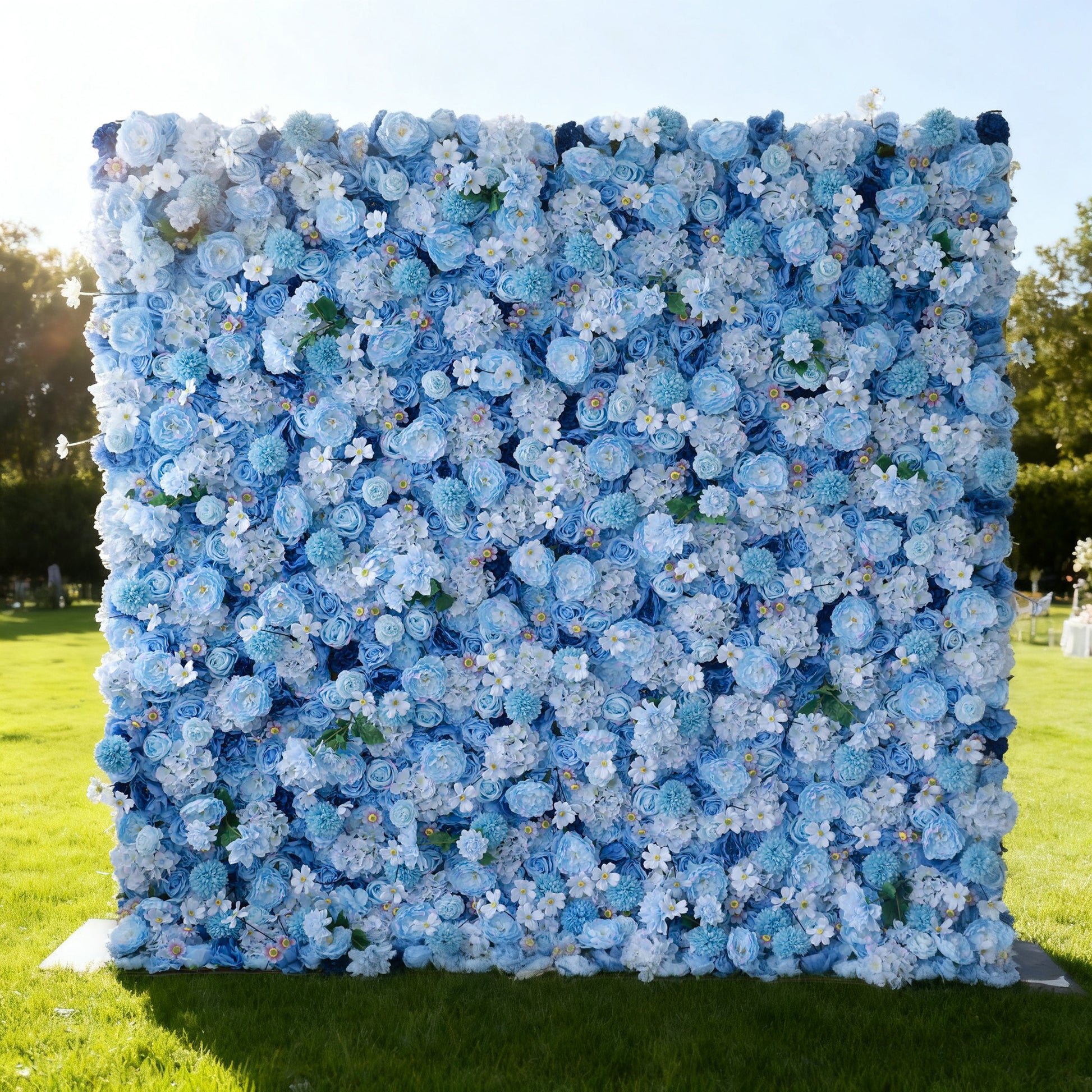 This image features a large floral wall outdoors on a lush green lawn. It's densely covered with artificial flowers in various shades of blue, from light sky blue to deeper hues, creating a soft and elegant color palette. Bathed in sunlight, with trees and a bright sky in the background, the floral wall exudes a serene and charming atmosphere, perfect for decorative use in events like weddings or photo shoots. Made by Valar Flowers.