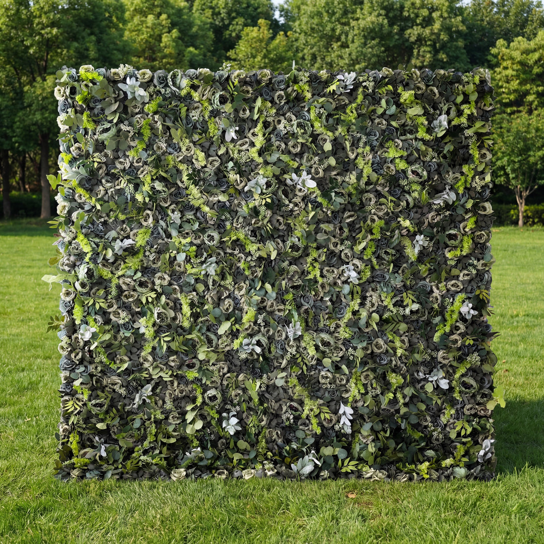 This lush, verdant outdoor backdrop features a dense, textured arrangement of artificial eucalyptus leaves, moss, and delicate filler foliage in varying shades of deep green, sage, and soft grey-green. Set on a sunlit, vibrant green lawn and framed by trees, its organic, monochromatic design exudes a modern, natural elegance, making it a sophisticated focal point for weddings, photoshoots, or upscale events seeking a fresh, earthy aesthetic.