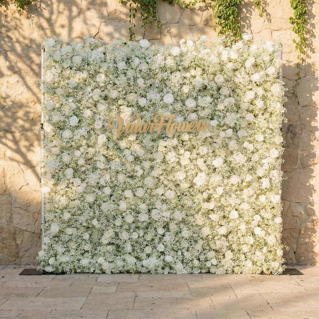 This Valar Flowers white flower wall backdrop is a lush, elegant tapestry of creamy white roses and delicate baby’s breath, creating a soft, cloud-like texture. Adorned with a custom gold brand sign, it’s set against a sunlit stone wall, offering a timeless, romantic focal point ideal for weddings, bridal showers, and refined photo opportunities.