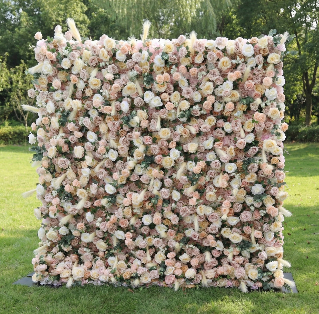This stunning outdoor floral wall from **Valar Flowers** is a lush tapestry of blush pink, creamy white, and soft peach blooms—including roses, peonies, and delicate filler flowers—interwoven with feathery pampas grass accents. Set on a sunlit green lawn framed by leafy trees, the dense, layered design creates a rich, three-dimensional texture, evoking a warm, romantic, and naturally elegant atmosphere ideal for weddings, photo backdrops, or elegant outdoor celebrations.