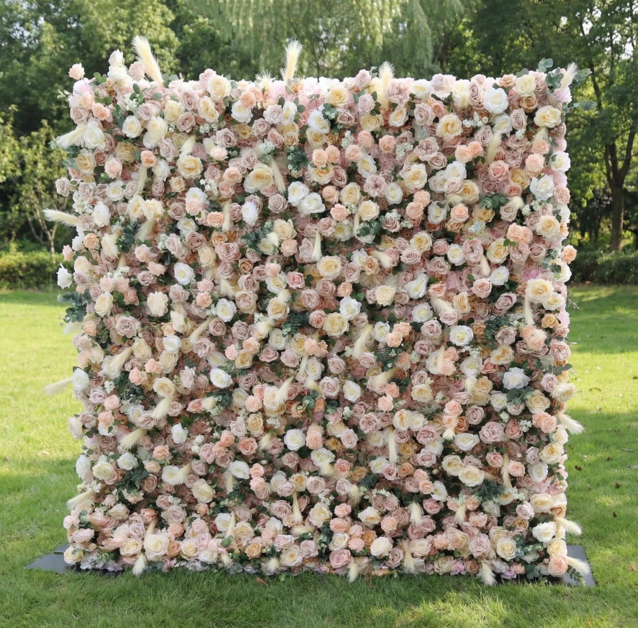 This stunning outdoor floral wall from **Valar Flowers** is a lush tapestry of blush pink, creamy white, and soft peach blooms—including roses, peonies, and delicate filler flowers—interwoven with feathery pampas grass accents. Set on a sunlit green lawn framed by leafy trees, the dense, layered design creates a rich, three-dimensional texture, evoking a warm, romantic, and naturally elegant atmosphere ideal for weddings, photo backdrops, or elegant outdoor celebrations.