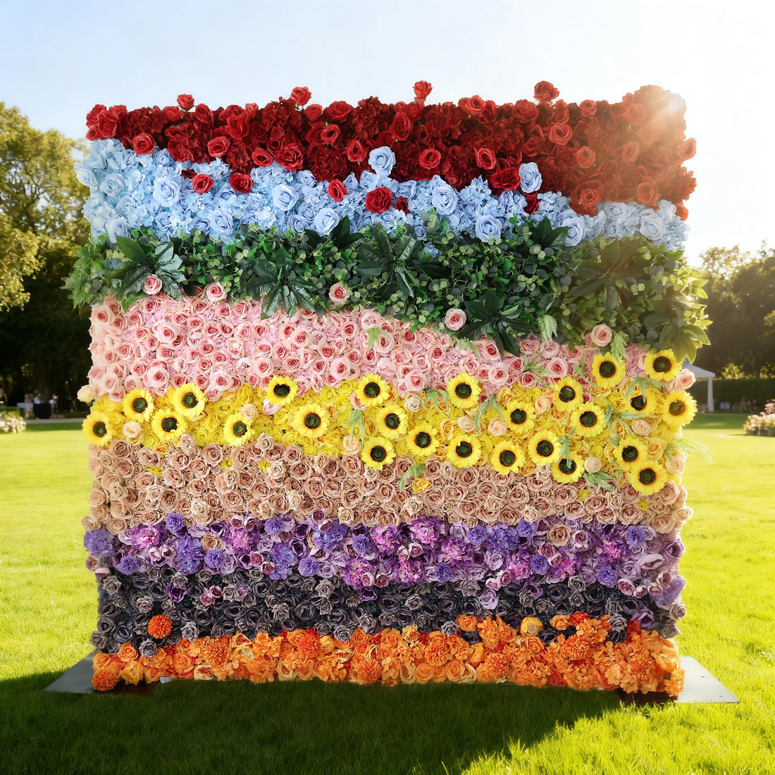 This image showcases a vibrant floral wall by Valar Flowers, set outdoors on a lush green lawn under bright sunlight. It features a stunning arrangement of artificial flowers in a spectrum of colors—red, blue, pink, yellow with sunflowers, purple, and orange—interspersed with green foliage. The layered, colorful design creates a lively and eye-catching display, perfect for adding a touch of natural beauty and charm to events or outdoor spaces.