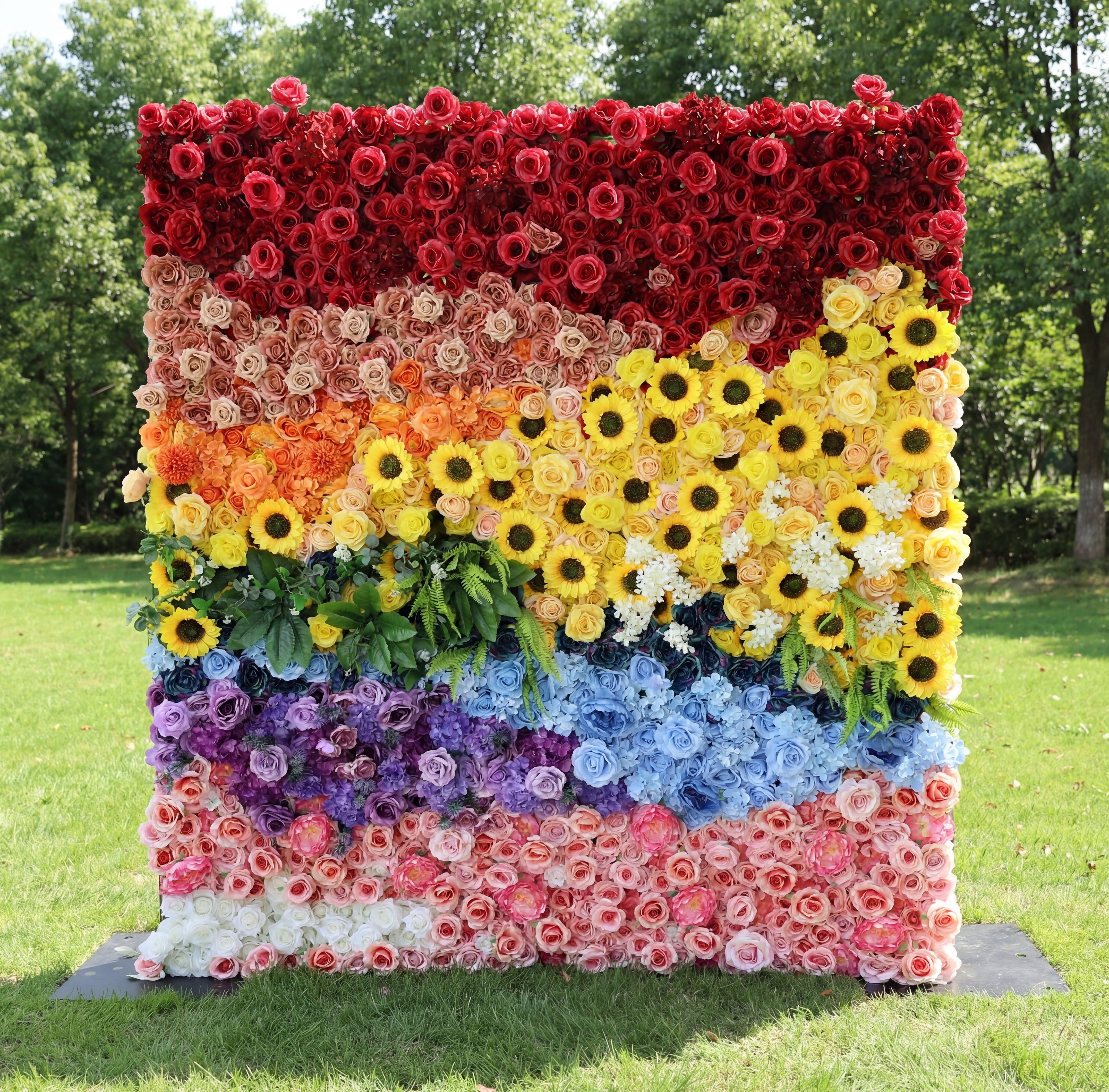 This vibrant outdoor floral backdrop from Valar Flowers features a bold, rainbow-inspired design, with distinct sections of rich red, warm peach, sunny yellow sunflowers, lush green foliage, dreamy blue and purple blooms, and soft pink and white roses. Set on a sunlit lawn framed by trees, its dense, textured arrangement of artificial flowers exudes joyful energy, making it a lively focal point for pride events, festivals, or colorful celebrations.