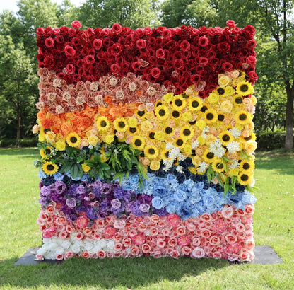 This vibrant outdoor floral backdrop from Valar Flowers features a bold, rainbow-inspired design, with distinct sections of rich red, warm peach, sunny yellow sunflowers, lush green foliage, dreamy blue and purple blooms, and soft pink and white roses. Set on a sunlit lawn framed by trees, its dense, textured arrangement of artificial flowers exudes joyful energy, making it a lively focal point for pride events, festivals, or colorful celebrations.