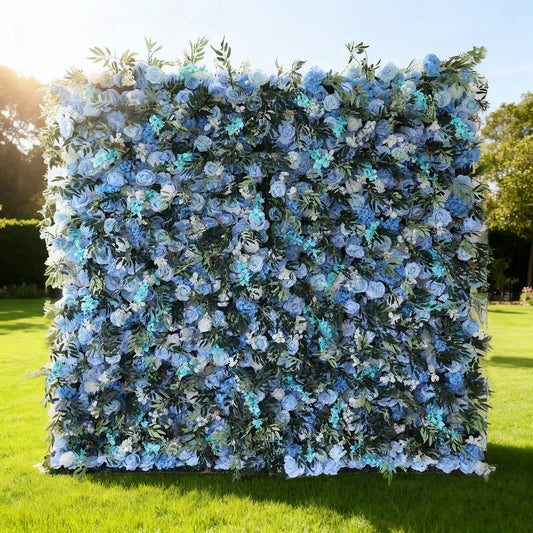 This image features a large floral wall outdoors on a lush green lawn. It's densely covered with artificial flowers in various shades of blue, from light sky blue to deeper hues, interspersed with green foliage. Bathed in sunlight, with trees and a bright sky in the background, the floral wall exudes a serene and elegant atmosphere, perfect for decorative use in events like weddings or photo shoots. Made by Valar Flowers.