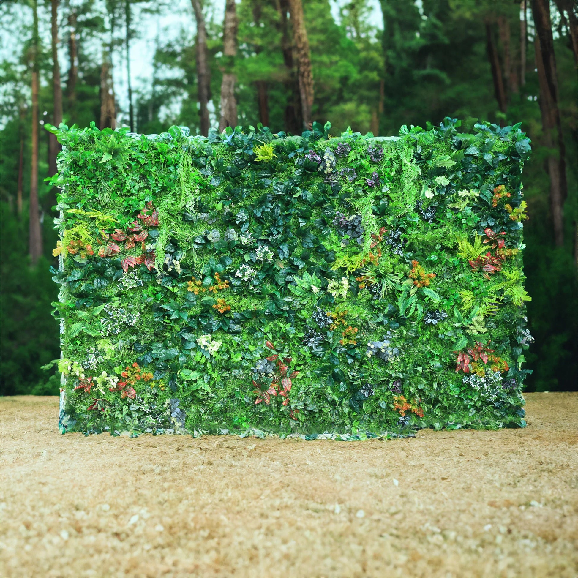 This image features a lush, artificial green wall set against a backdrop of a forested area. The green wall is densely packed with a variety of faux plants, including different shades of green foliage, some with variegated patterns, and splashes of red, orange, and white for added color. The wall appears full and vibrant, creating a natural - looking and inviting scene, ideal for events or as a decorative partition. 
