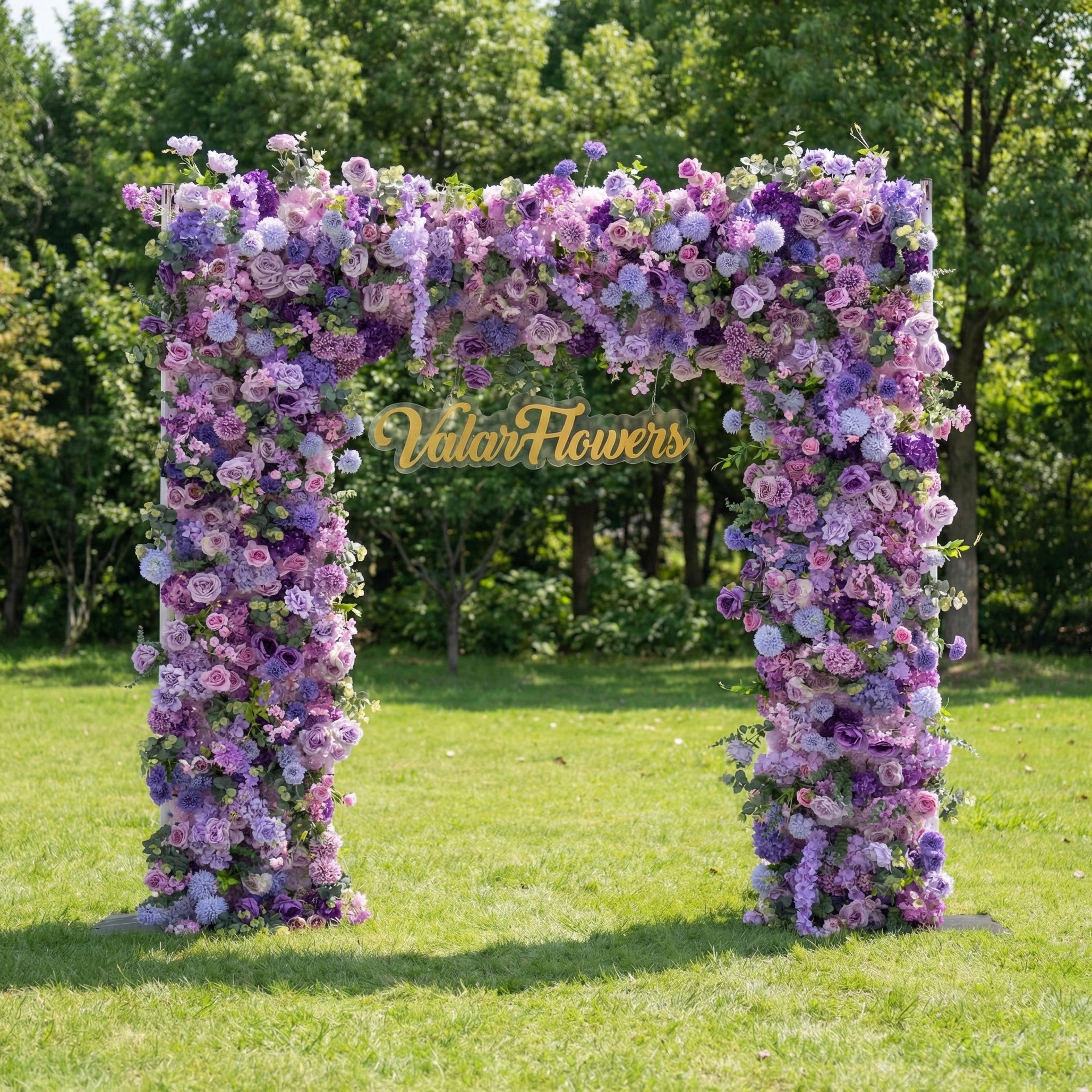 This enchanting floral arch from Valar Flowers is densely adorned with artificial blooms in a dreamy palette of soft lavender, lilac, and blush pink, layered with delicate filler flowers for a lush, three-dimensional effect. Set on a sunlit green lawn framed by leafy trees, its romantic, ethereal design exudes timeless elegance and whimsy, making it a stunning focal point for weddings, garden parties, or photoshoots seeking a soft, romantic aesthetic.