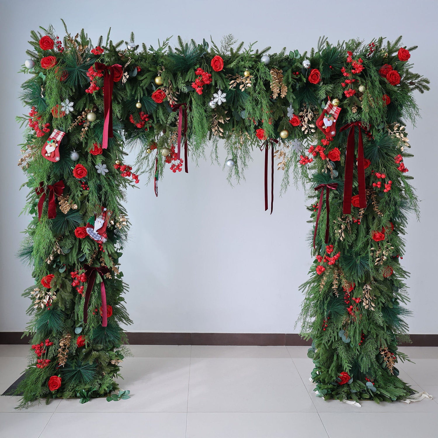 This image shows a festive Christmas arch adorned with lush green foliage. It features vibrant red roses, berries, and decorative elements like small stockings, ribbons, and baubles in gold, silver, and red. The arch, set against a plain light - colored wall and tiled floor, exudes a warm, holiday charm, perfect for Christmas celebrations and creating a joyful, festive atmosphere.