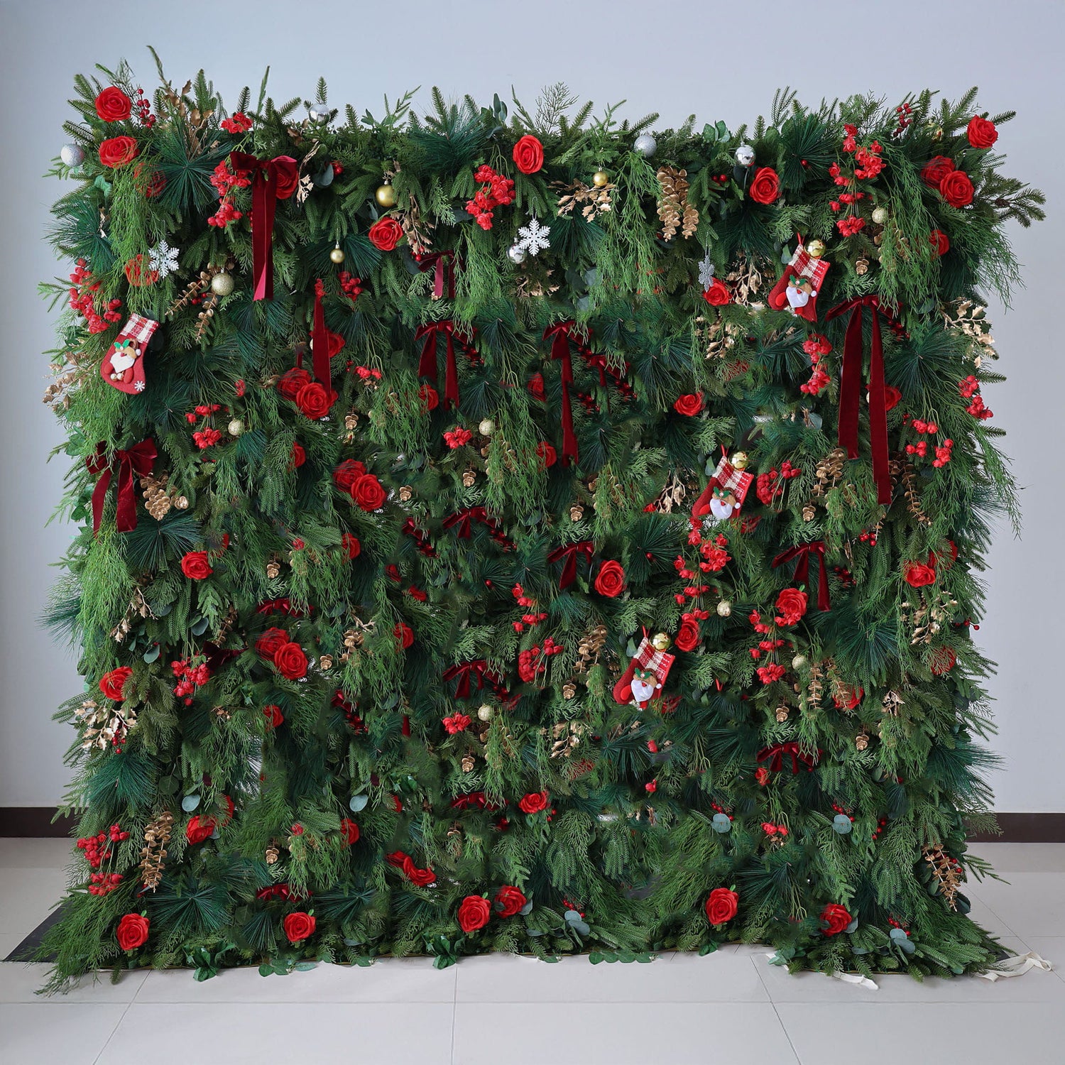 This image displays a large, festive Christmas - themed floral wall. It is richly decorated with lush green foliage, vibrant red roses, and various Christmas ornaments such as small stockings, snowflakes, and baubles. Deep red ribbons are also incorporated, adding to the holiday charm. Set against a plain light - colored wall and a tiled floor, the wall exudes a warm and joyful Christmas atmosphere, perfect for holiday decorations.