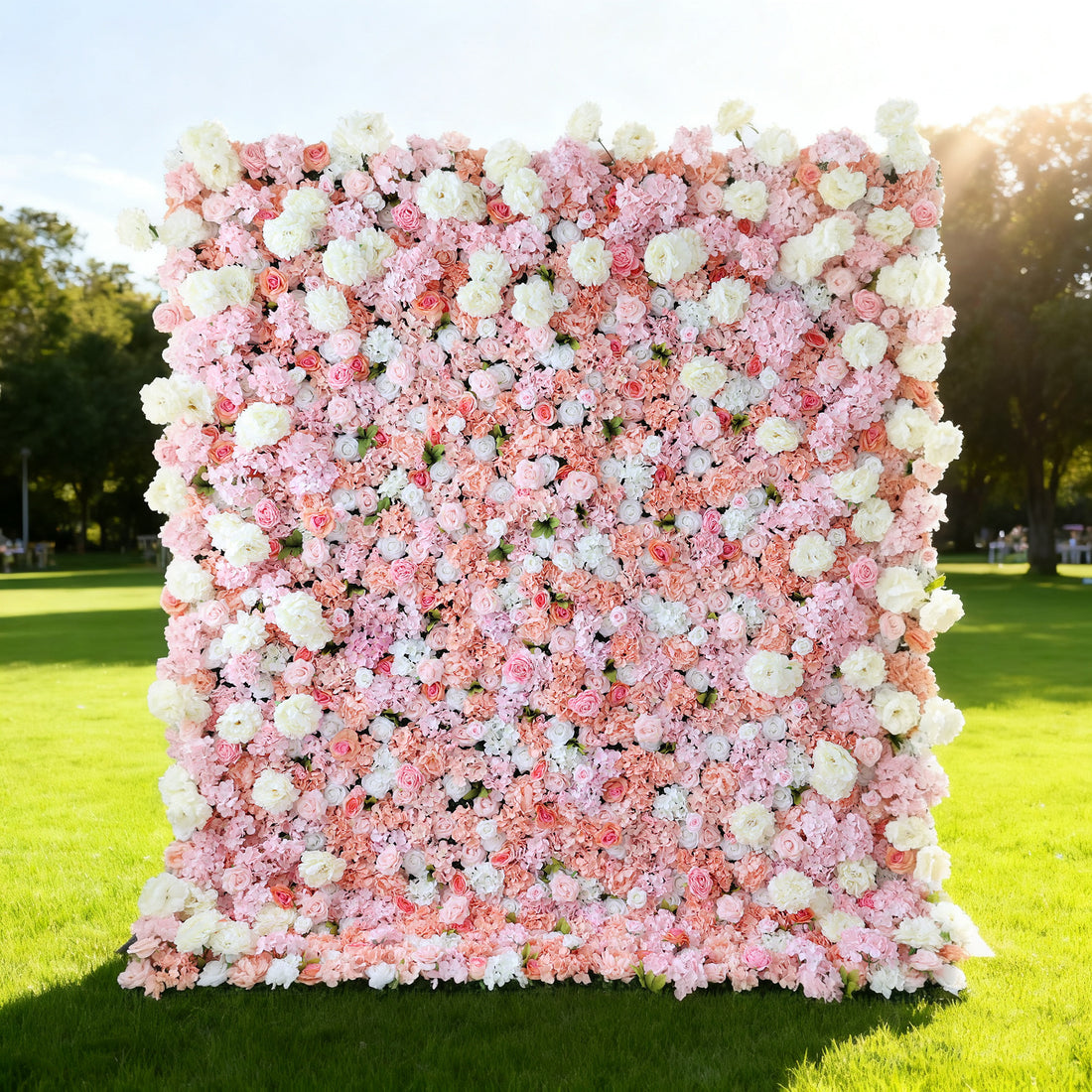 This image showcases a lush floral backdrop by ValarFlowers, set outdoors on a vibrant lawn. It features a dense arrangement of artificial flowers in shades of pink, peach, and white, creating a luxurious, textured display. Bathed in sunlight with a scenic park in the background, it exudes elegance, perfect for adding a nature-inspired, sophisticated element to outdoor events.