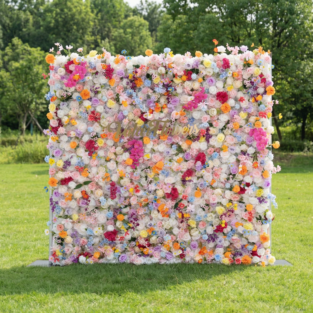 This vibrant outdoor floral backdrop stands on a sunlit green lawn, framed by lush trees. It is a dense, joyful arrangement of artificial blooms in a rainbow of soft pastels and bright pops of color, including pinks, oranges, yellows, and blues. The multi-tonal design creates a cheerful, eye-catching focal point, perfect for weddings, parties, or photoshoots, blending whimsical charm with a lively, garden-inspired aesthetic.