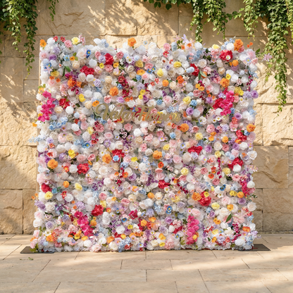 This is a stunning, dense artificial flower wall backdrop from Valar Flowers, bursting with a vibrant, rainbow-hued mix of lifelike blooms. Set against a sunlit stone wall with trailing ivy, it features creamy white roses, soft pink peonies, sunny yellow poppies, vivid orange carnations, sky-blue delphiniums, deep magenta orchids, and delicate filler flowers, creating a lush, romantic focal point perfect for weddings, photo booths, and luxury event decor.