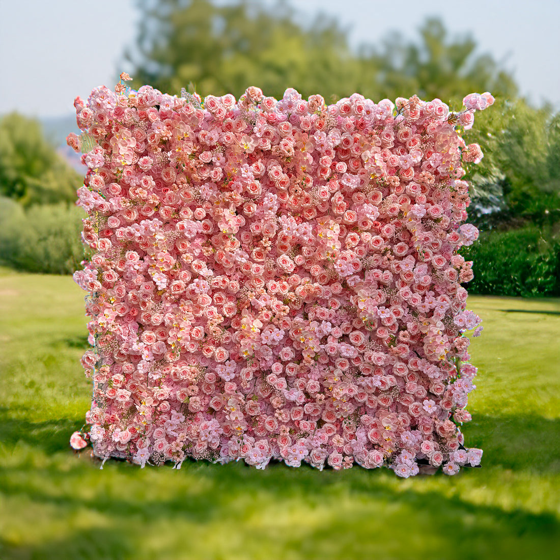 This photo showcases a large, square floral backdrop densely covered with an abundance of soft pink roses and other delicate pink flowers. Set against a lush green grassy field with blurred greenery in the background, it exudes a serene and romantic ambiance. Ideal for events like garden weddings or elegant outdoor parties, it creates a dreamy and picturesque setting.