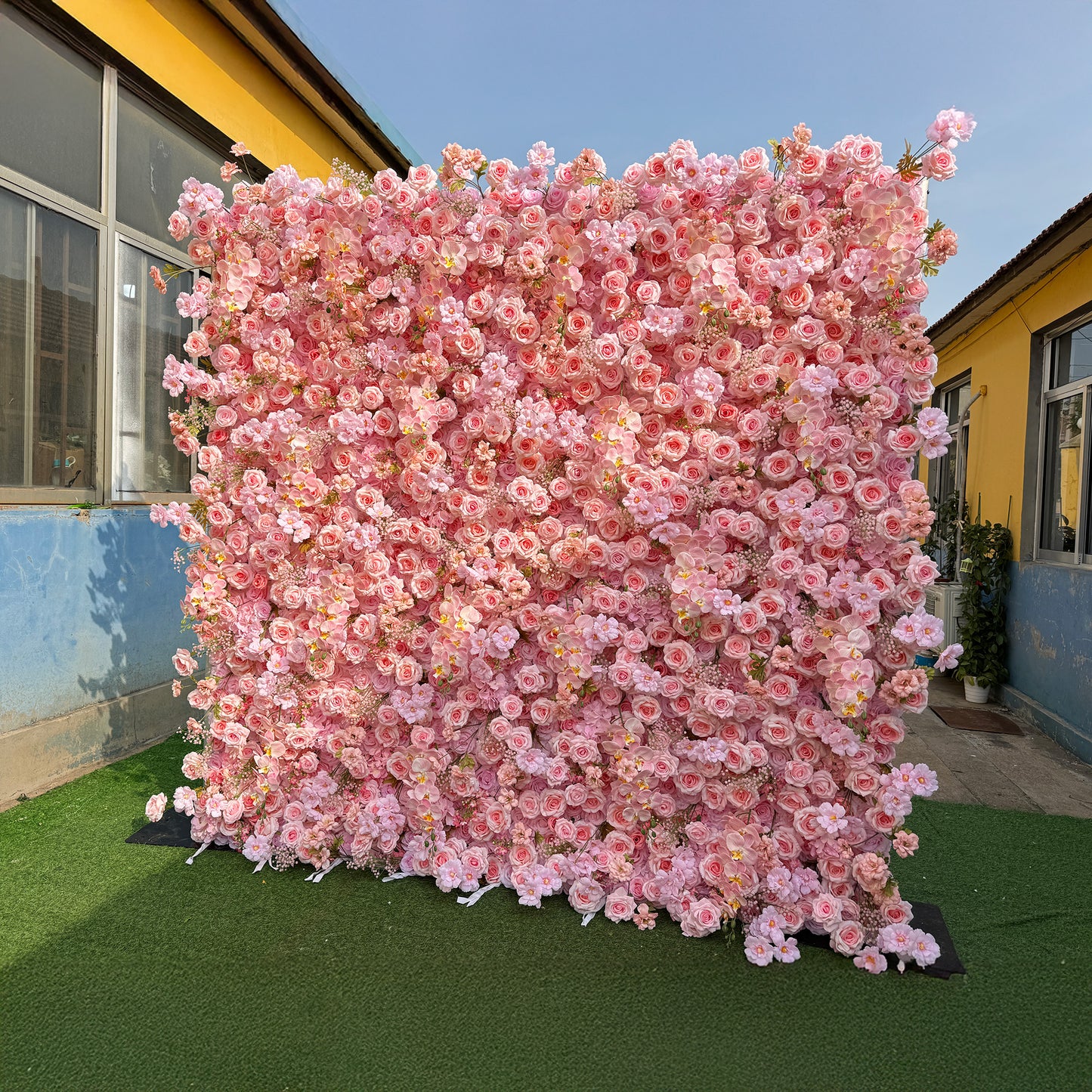 This photo showcases a large, square floral backdrop densely packed with soft pink roses and other delicate pink flowers. Set against a backdrop of a yellow and blue building, it exudes a charming and romantic atmosphere. The lush arrangement creates a visually stunning display, perfect for events like weddings, engagement parties, or high - end outdoor gatherings. 