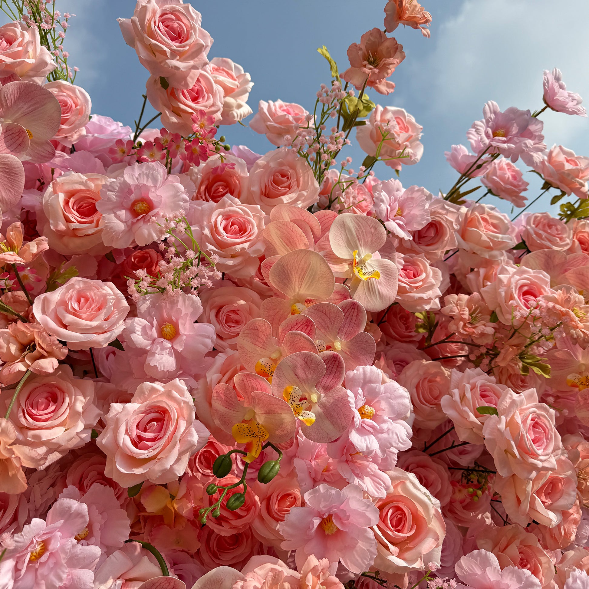 This photo showcases a lush and abundant floral arrangement filled with soft pink roses, delicate pink orchids, and small pink blossoms. The pastel - colored flowers create a gentle and dreamy atmosphere, set against a backdrop of a partly cloudy blue sky. The dense cluster of blooms exudes a sense of romance and elegance, perfect for events like weddings or high - end garden parties. 