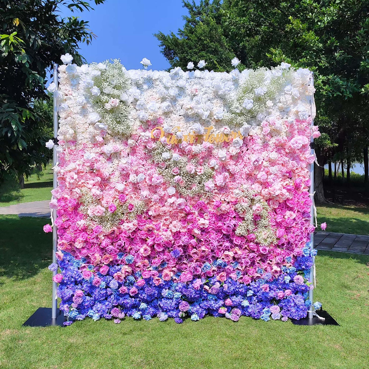 This photo features a lush floral wall with a gradient of white, pink, and blue. Composed of roses and hydrangeas, with baby's breath adding texture. Set outdoors on green grass with trees in the background, it has a dreamy, romantic style, ideal for weddings or photo - booth backdrops. 